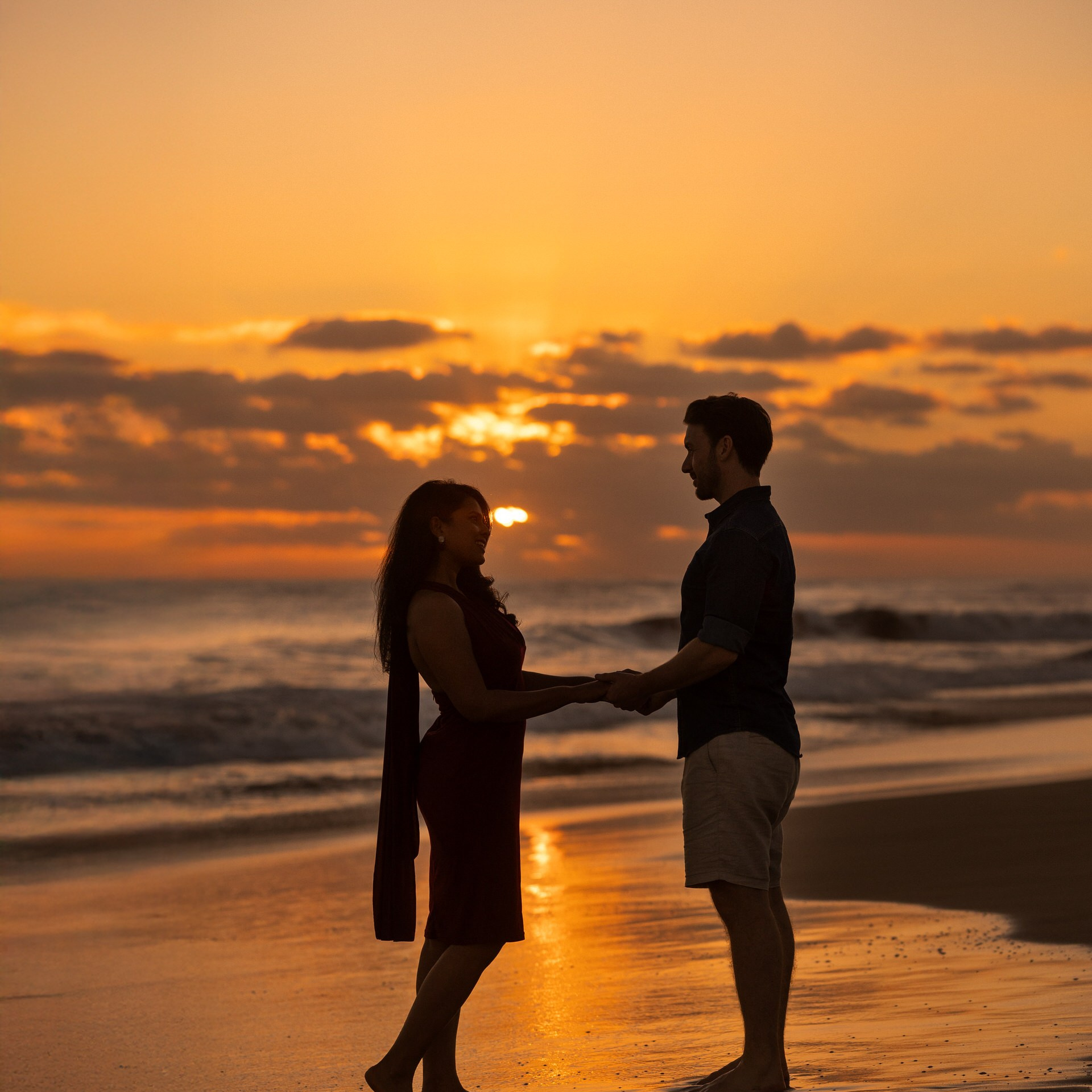 A couple stands barefoot on the beach at sunset, holding hands and facing each other with the ocean and orange sky in the background at Faro Maspalomas