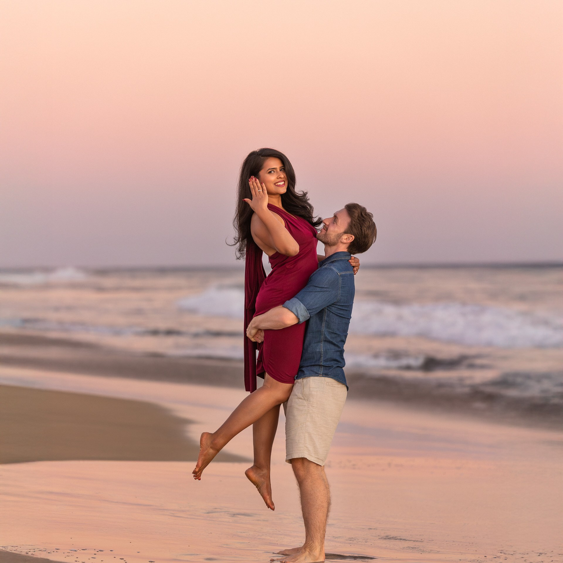 A man lifts a woman in a red dress while standing on a beach Maspalomas at sunset