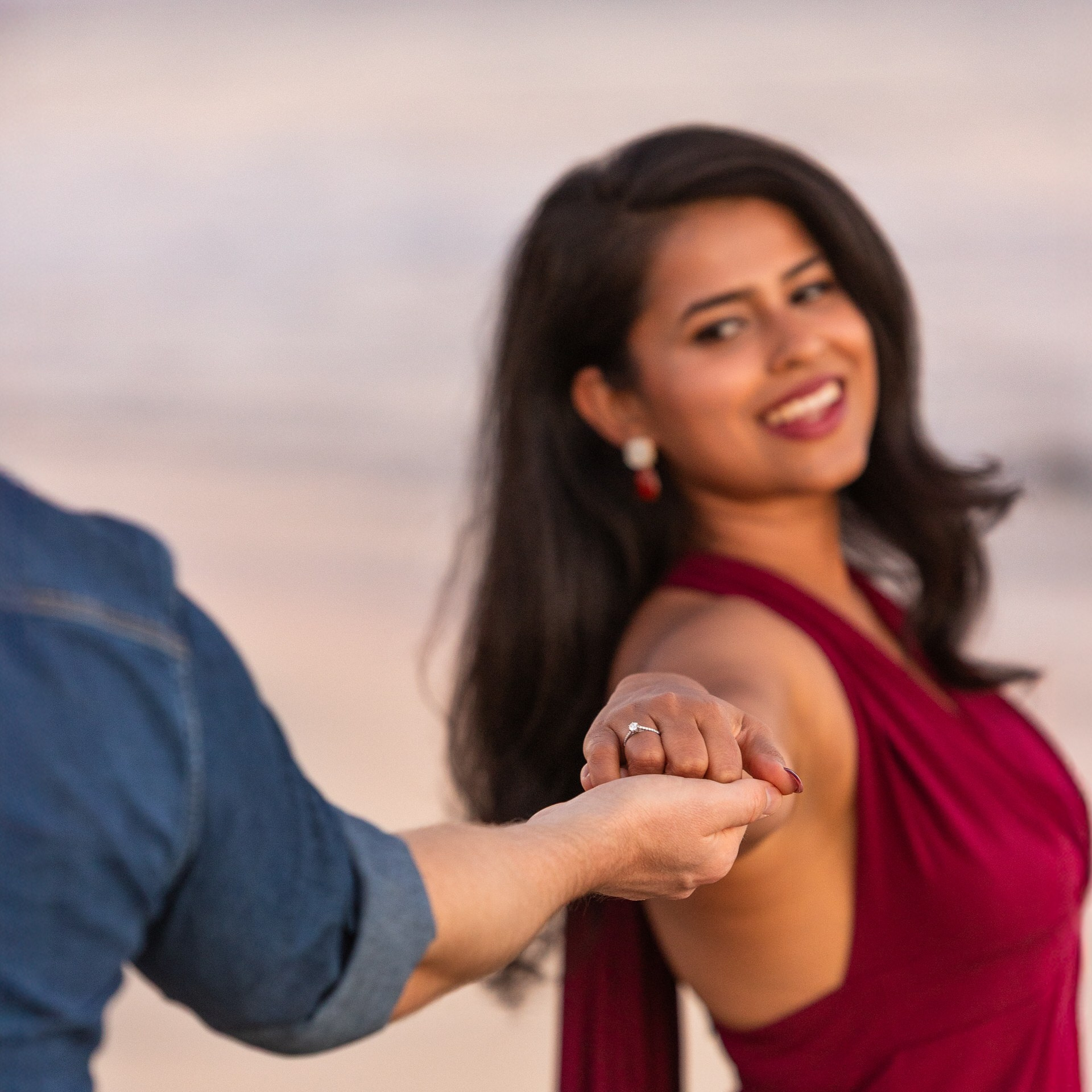 A woman in a burgundy dress smiles while holding hands with someone, standing outdoors near a body of water.