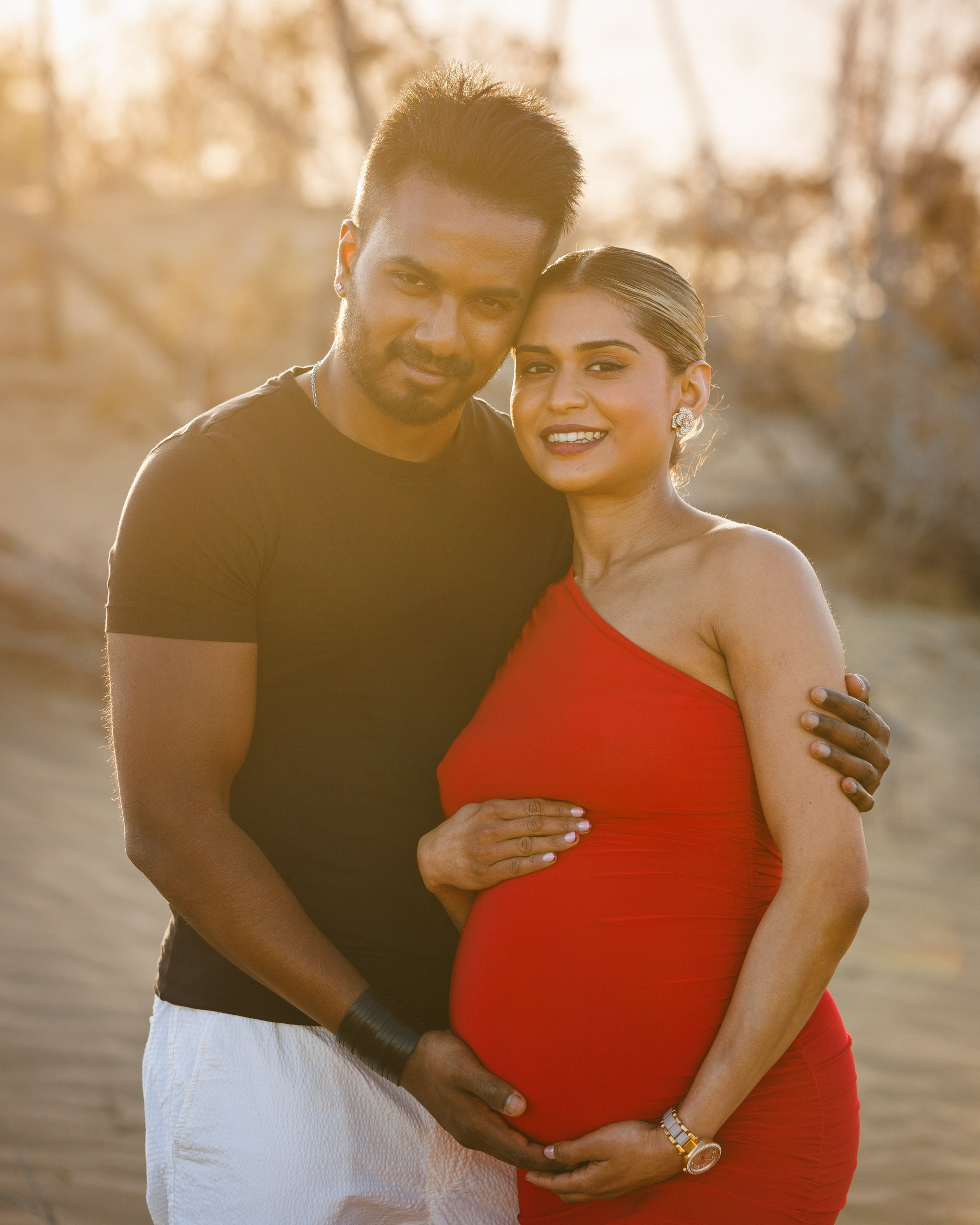 Maspalomas beach A couple stands outdoors, with the man in a black shirt and white pants, and the woman in a red dress holding her pregnant belly, both smiling at the camera.