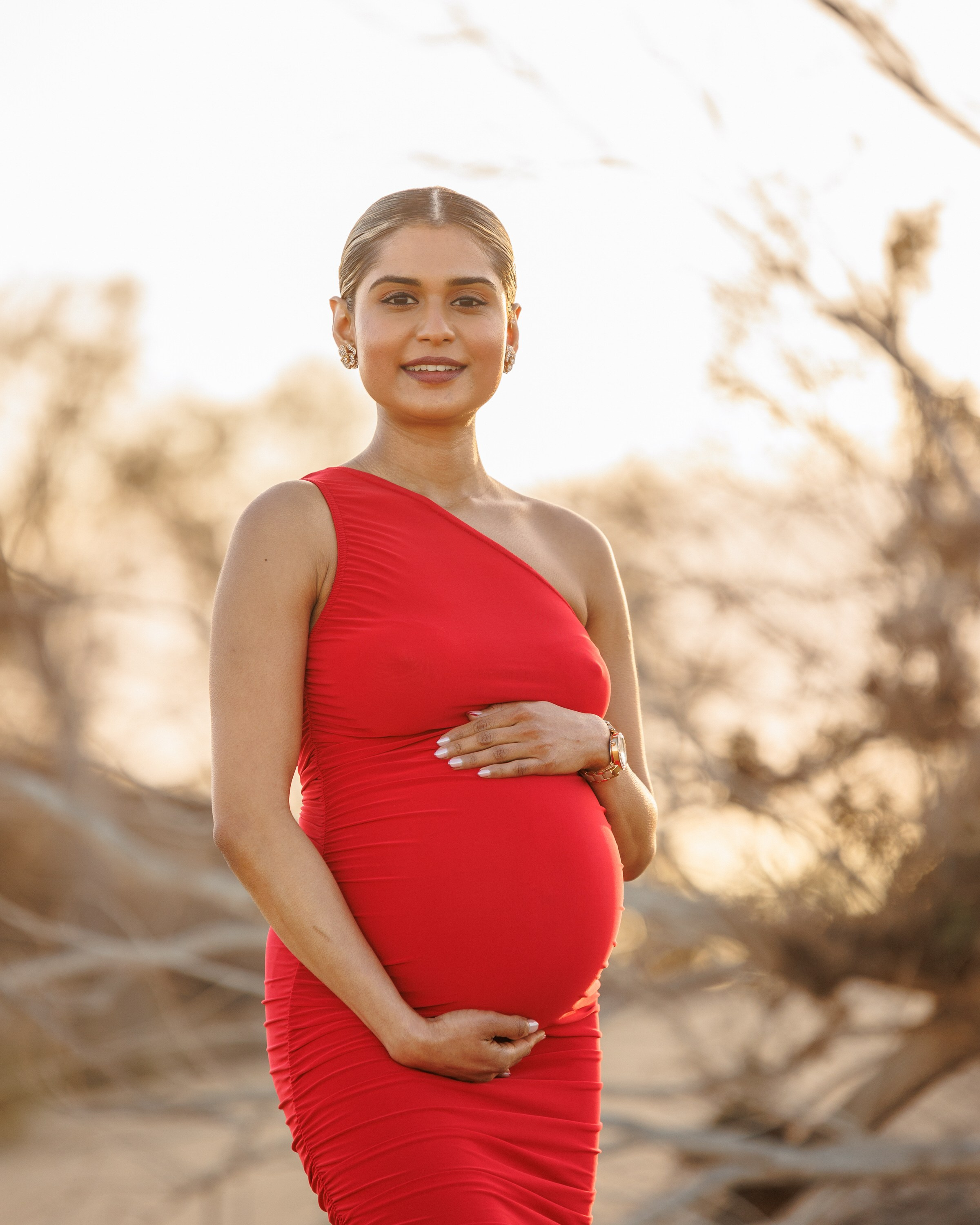 Meloneras Gran Canaria A pregnant woman in a red, one-shoulder dress stands outdoors, holding her belly with both hands and smiling at the camera.