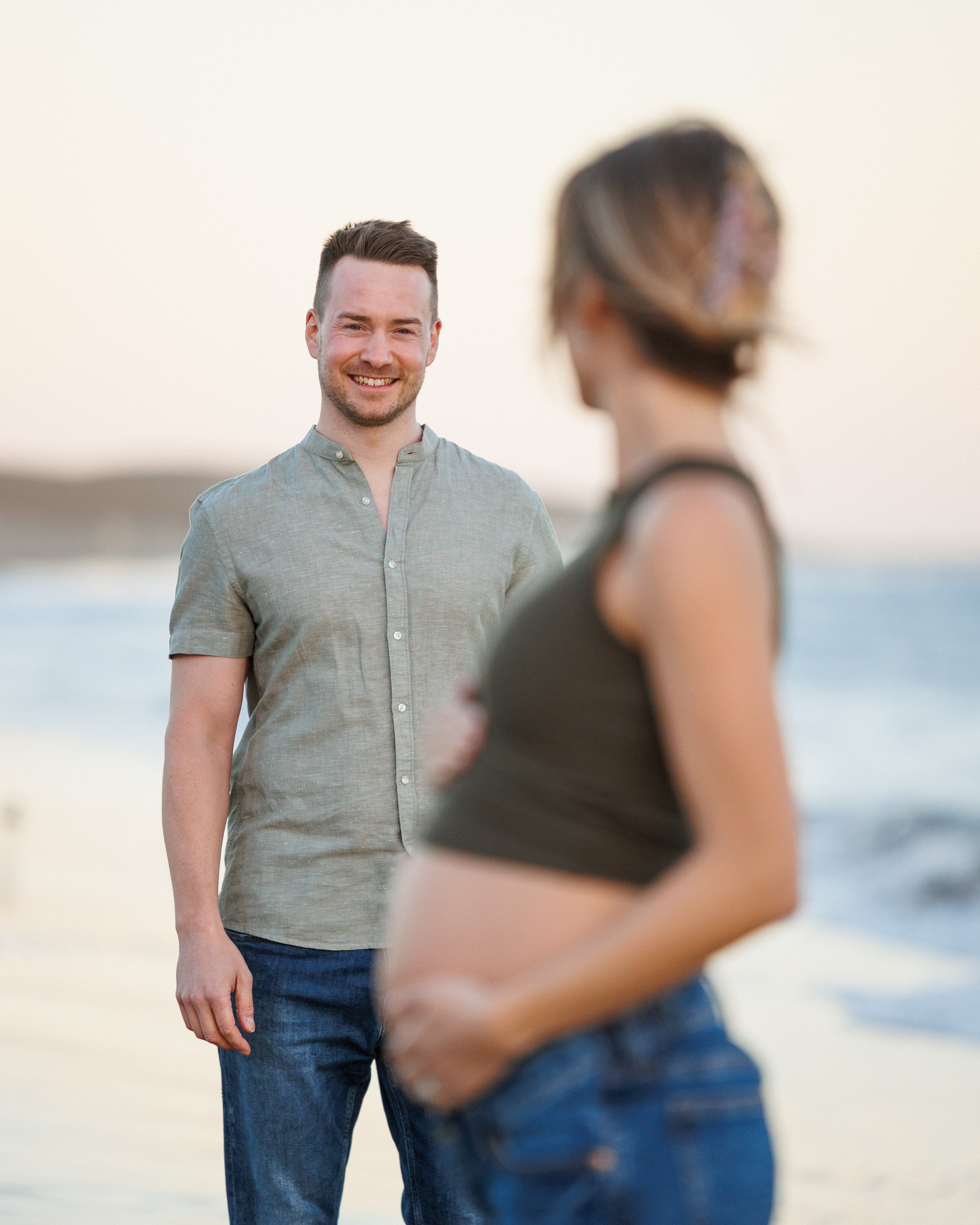 Maspalomas Gran Canaria A man stands smiling on a beach while a pregnant woman in the foreground faces him, holding her belly.