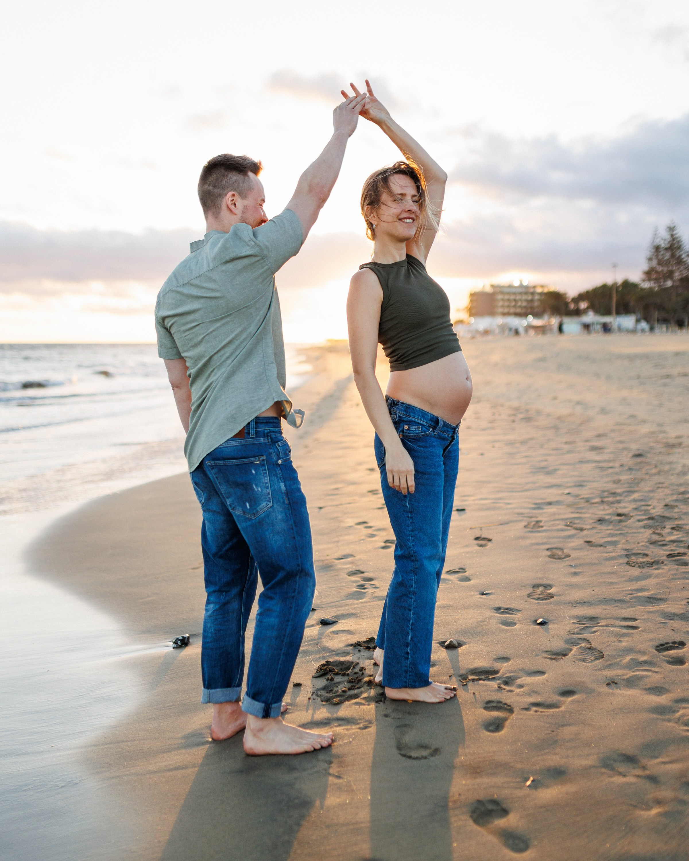 Maspalomas Gran Canaria A barefoot couple dances on a sandy beach at sunset. The woman is visibly pregnant, and both are dressed casually in jeans and shirts.