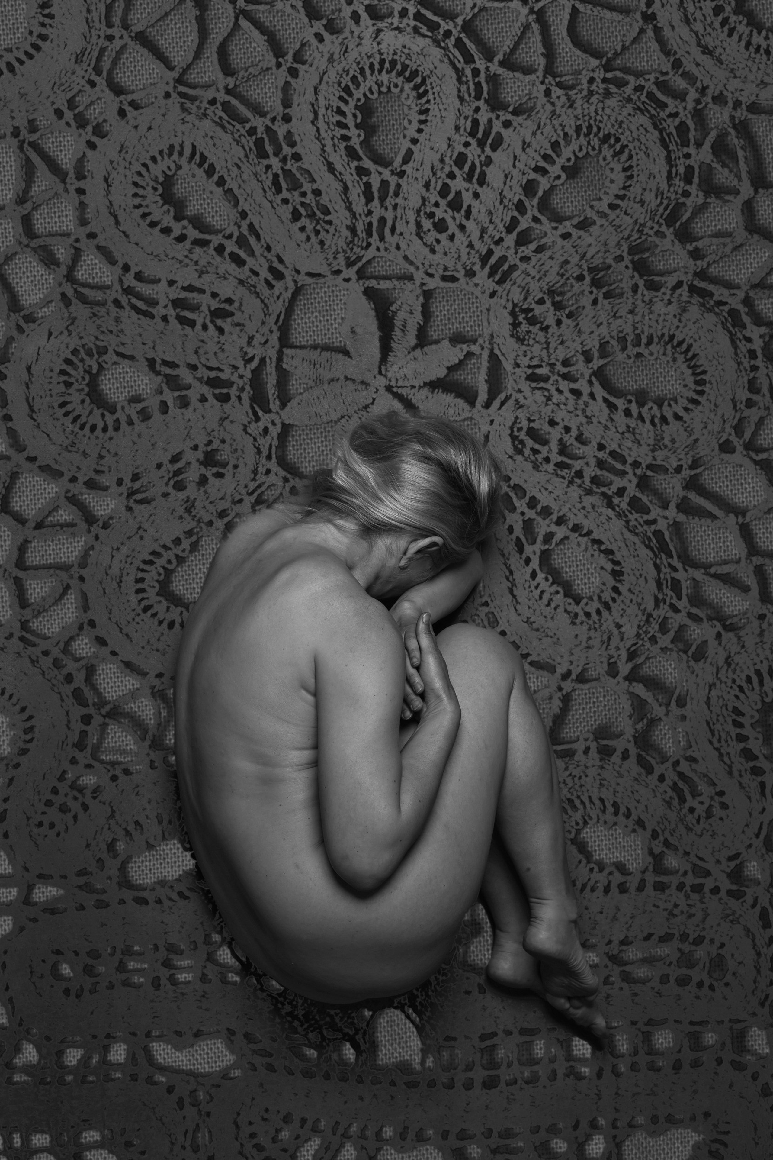 Black-and-white fine art photograph of a nude woman curled in fetal position on a large lace textile, symbolizing rebirth, vulnerability, and ancestral memory – photography by Maria Tulchinskaya.