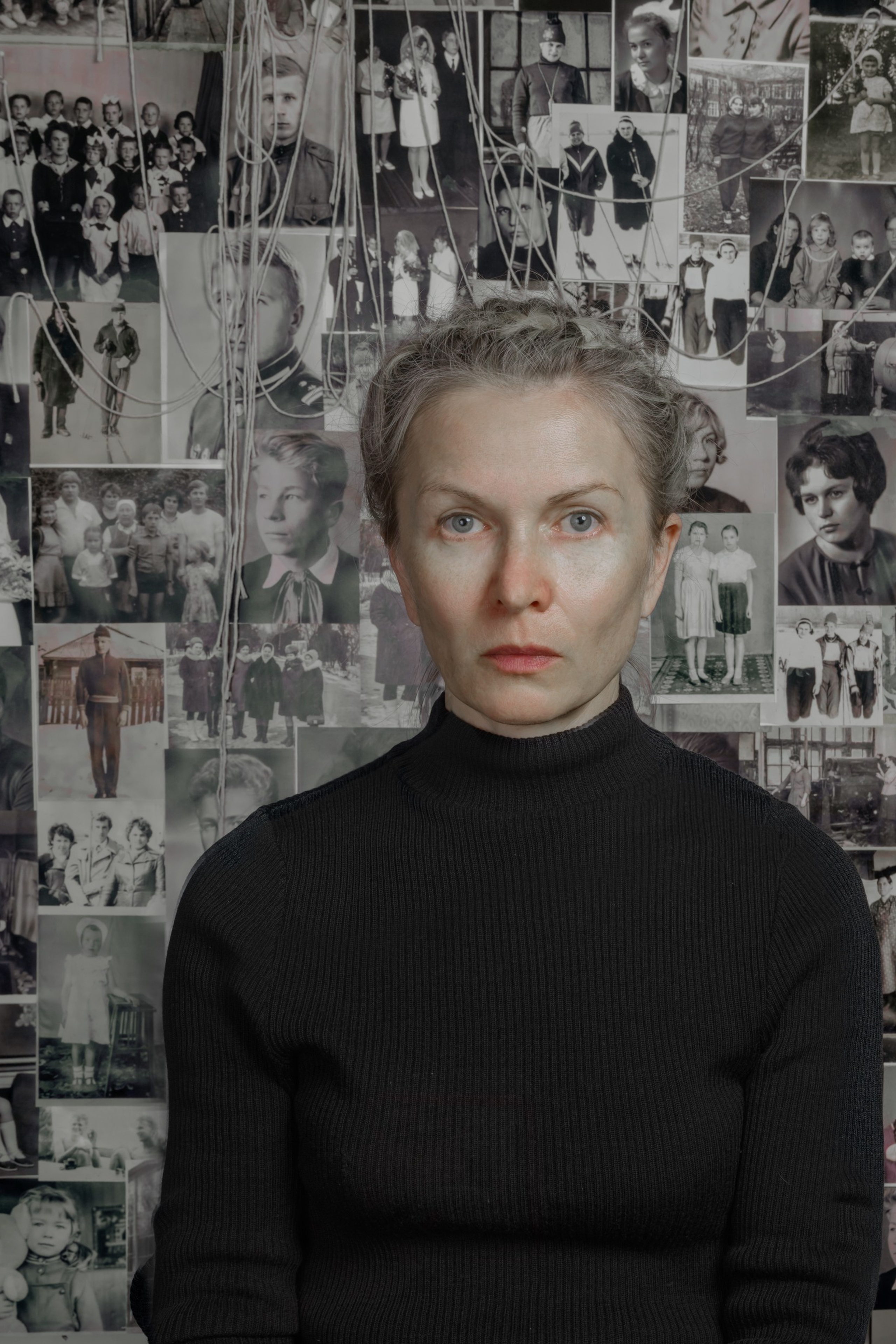 Portrait of a woman in black in front of a collage of old family photographs connected with threads, symbolizing ancestral memory, fragmented identity, and generational continuity – fine art photography by Maria Tulchinskaya.