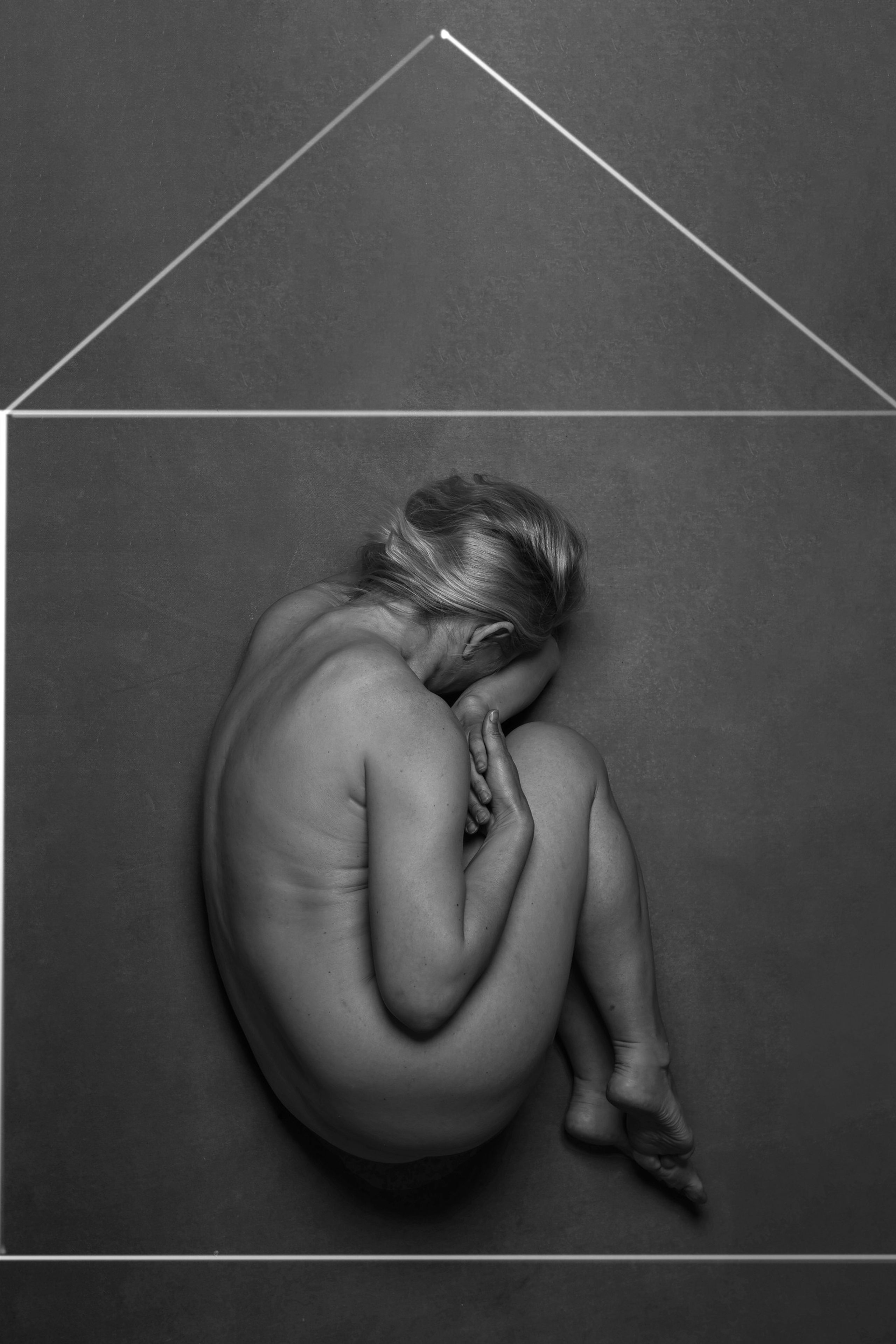 This black and white photograph of a naked woman curled up in a fetal position inside a visually drawn house
