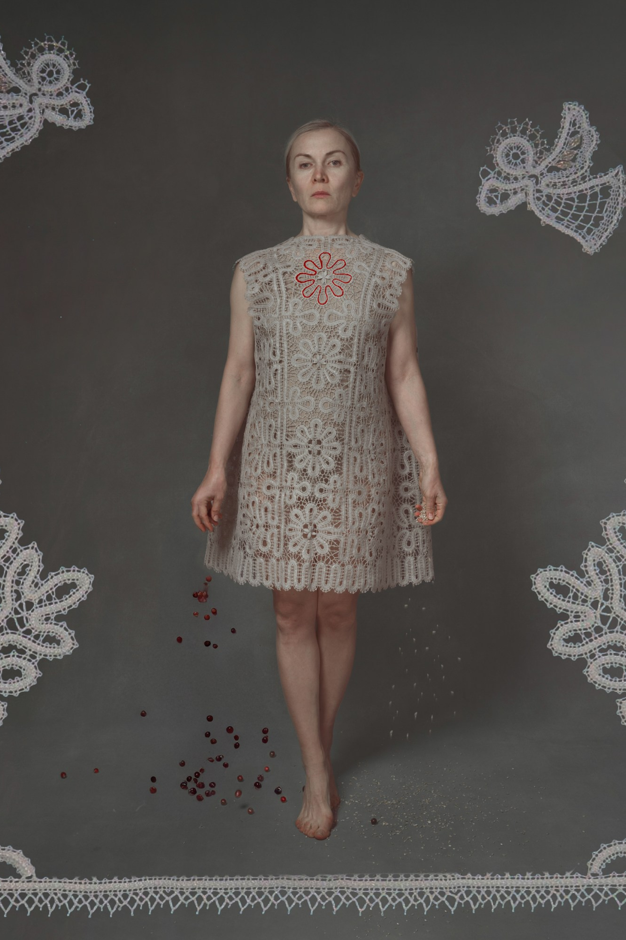 Symbolic portrait of a woman standing barefoot in a lace garden with embroidered heart motif, surrounded by lace angels and trees, evoking themes of memory, sacred femininity, and ancestral ritual – fine art photography by Maria Tulchinskaya.