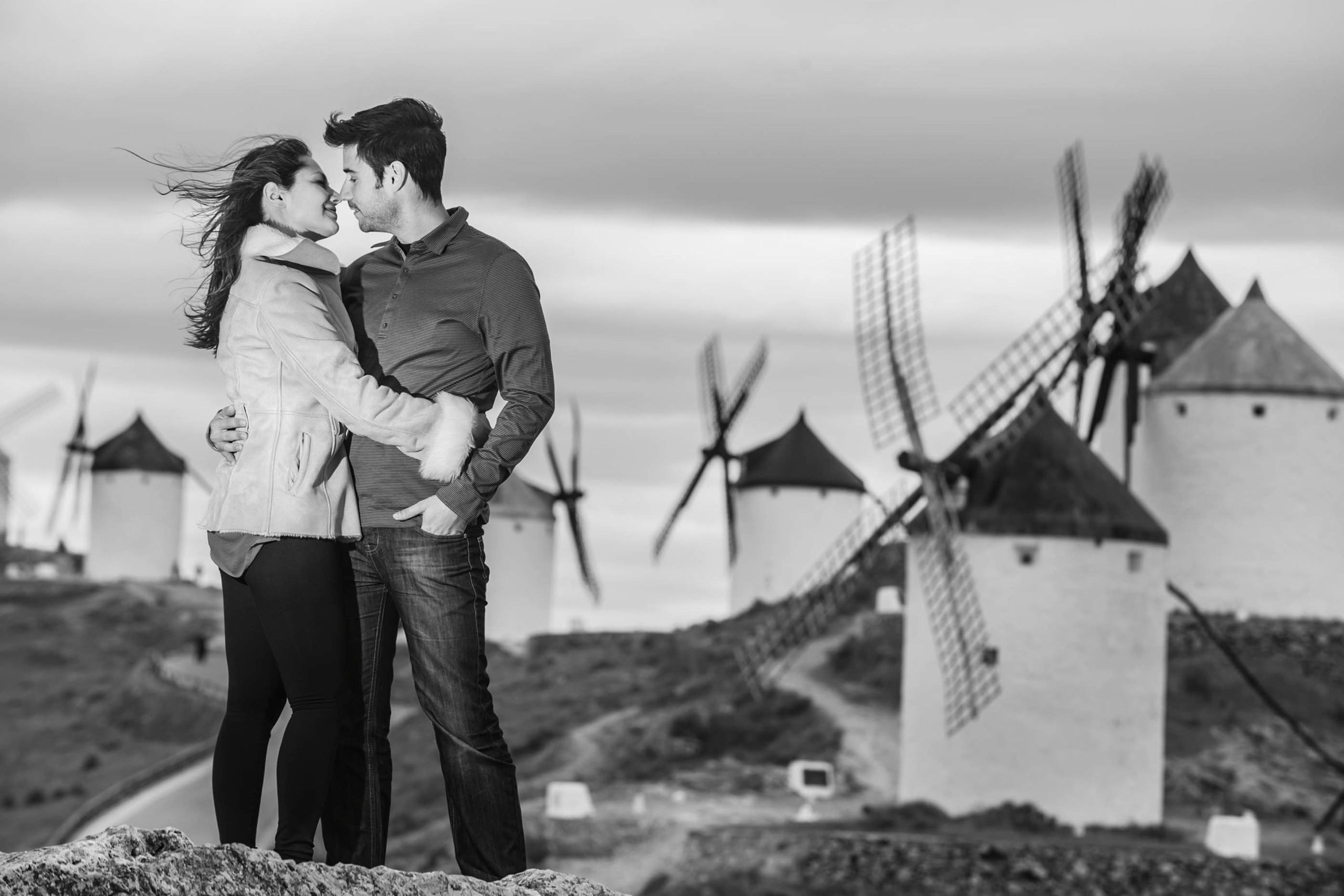A couple facing each other with traditional windmills in the background on a cloudy day in Toledo, Spain