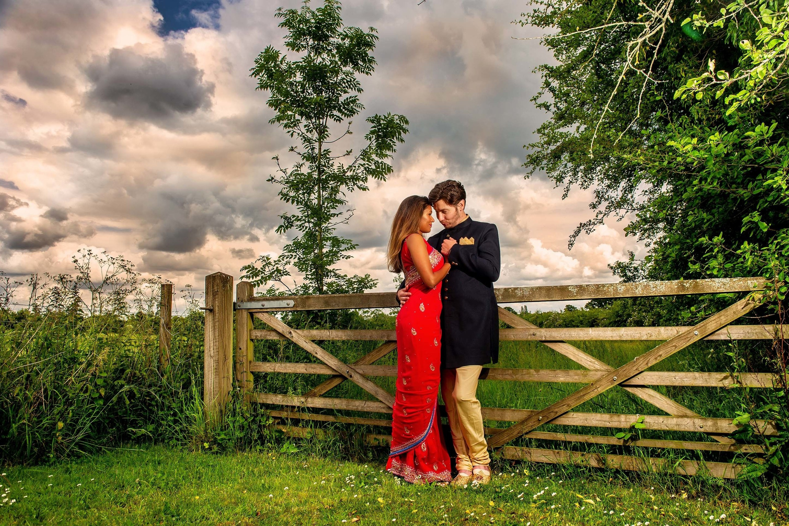 A couple dressed in traditional attire embraces a wooden gate with a dramatic sky in the background during a countryside engagement photoshoot.