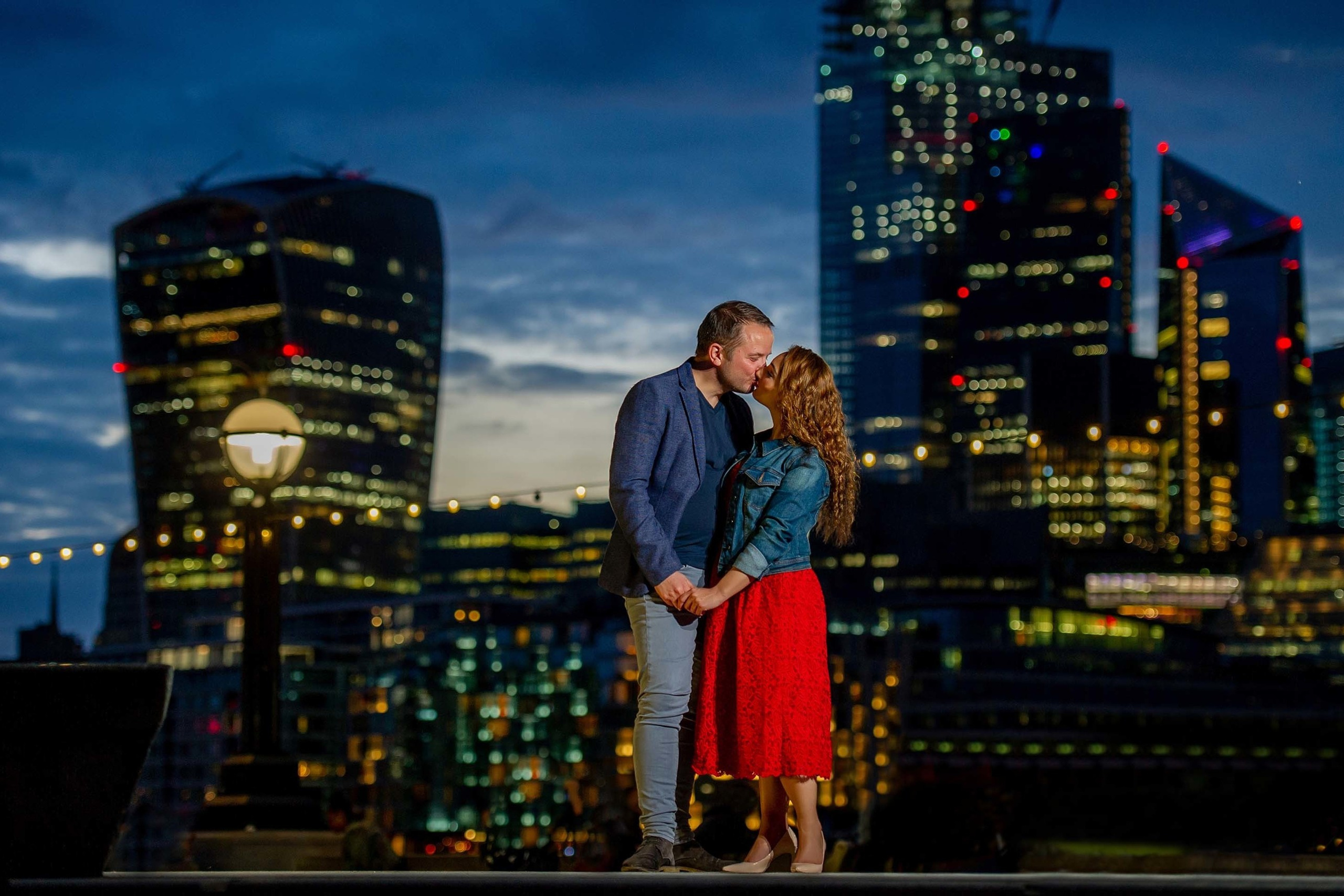 A couple kissing under string lights with London skyscrapers lit up at night.