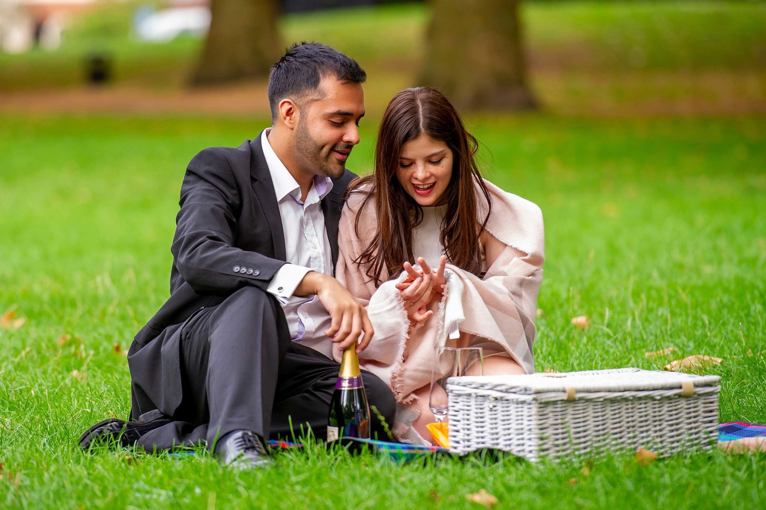 Man proposing to his partner with a picnic basket and champagne on the grass in a romantic park setting