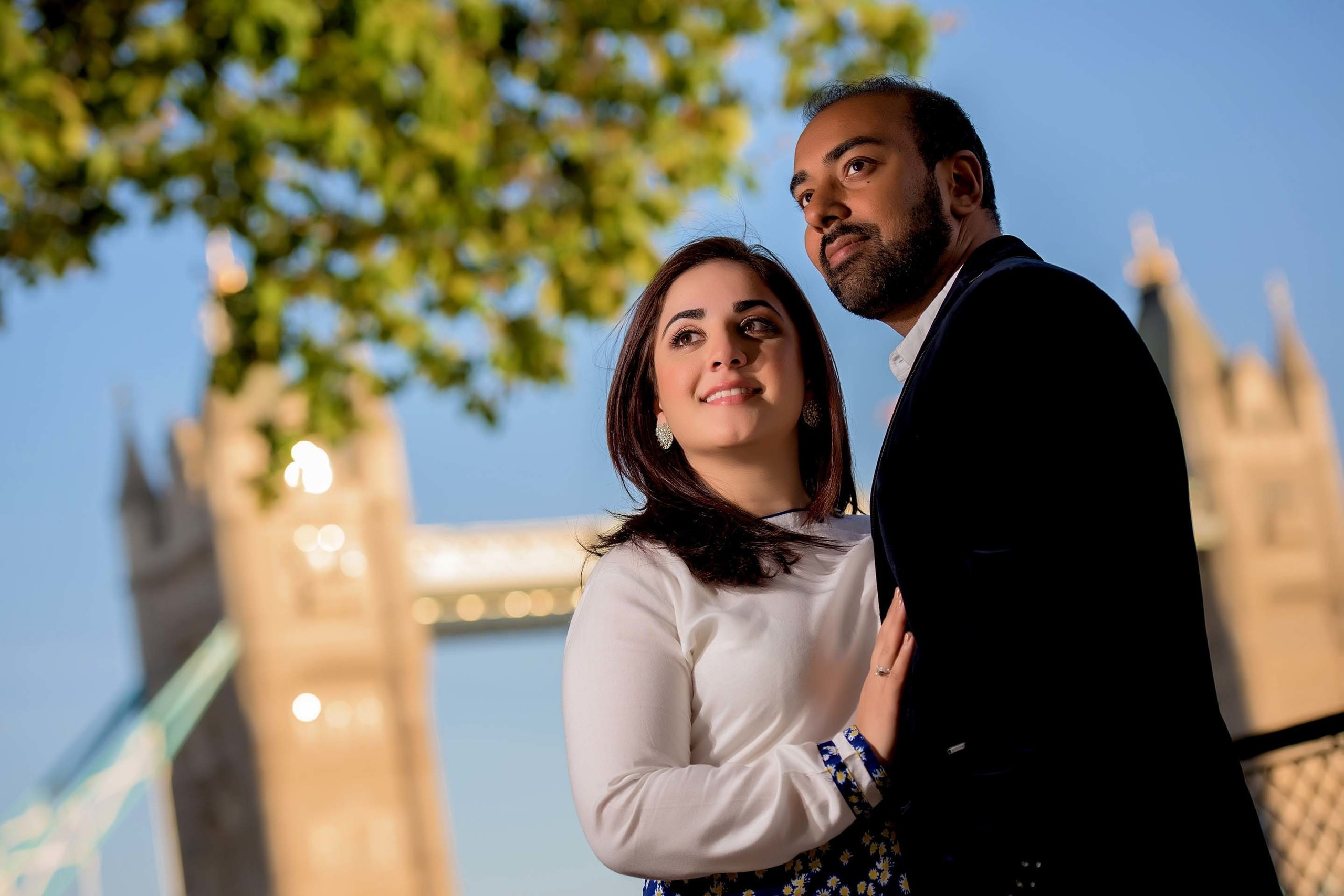 Smiling couple posing in front of Tower Bridge in London during engagement shoot under a clear blue sky