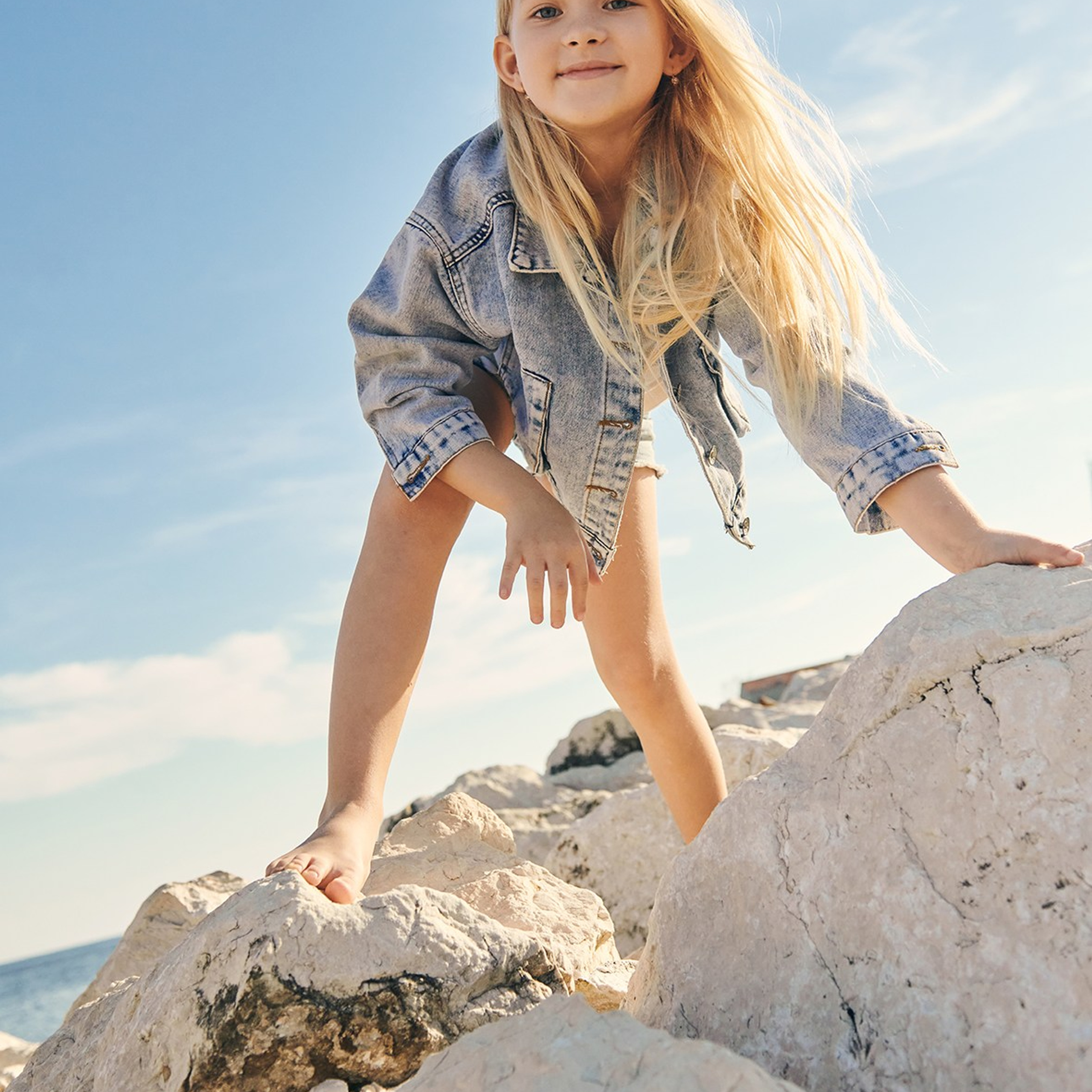 Fashion portrait of a blonde long haired girl in a denim dress claiming a rock during a children’s photoshoot