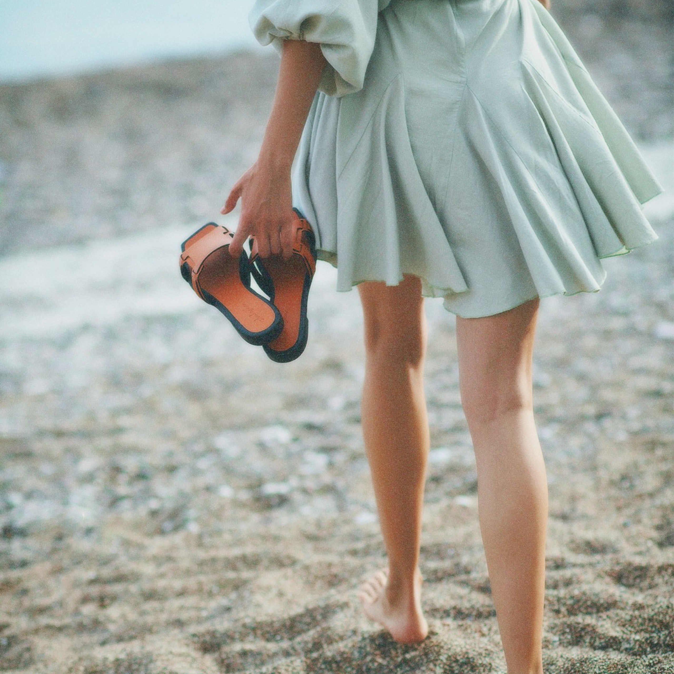 Fashion portrait of a long-legged slender woman carrying brown leather sandals by the sea