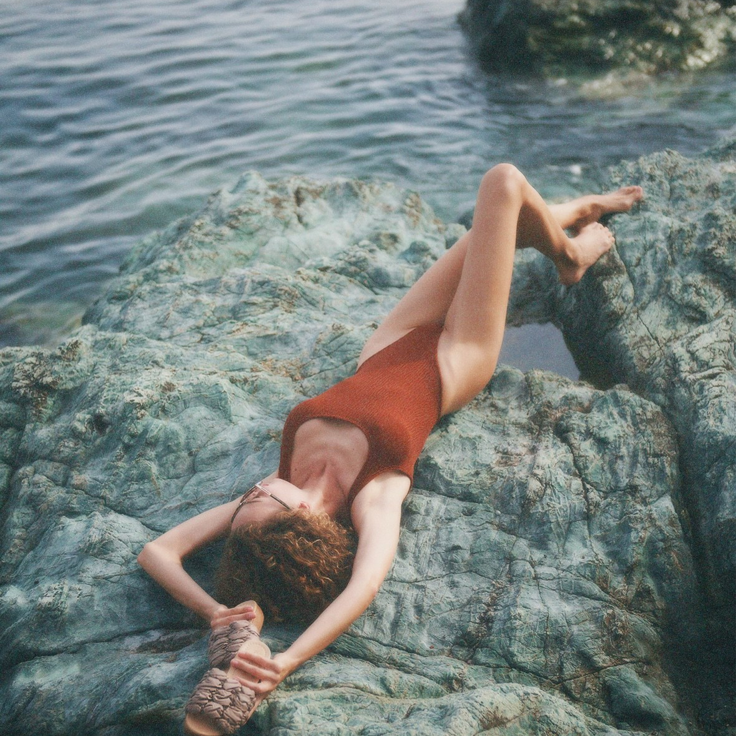 Fashion photoshoot of a woman in a brown swimsuit posing on a rock by the sea, advertising stylish beach footwear
