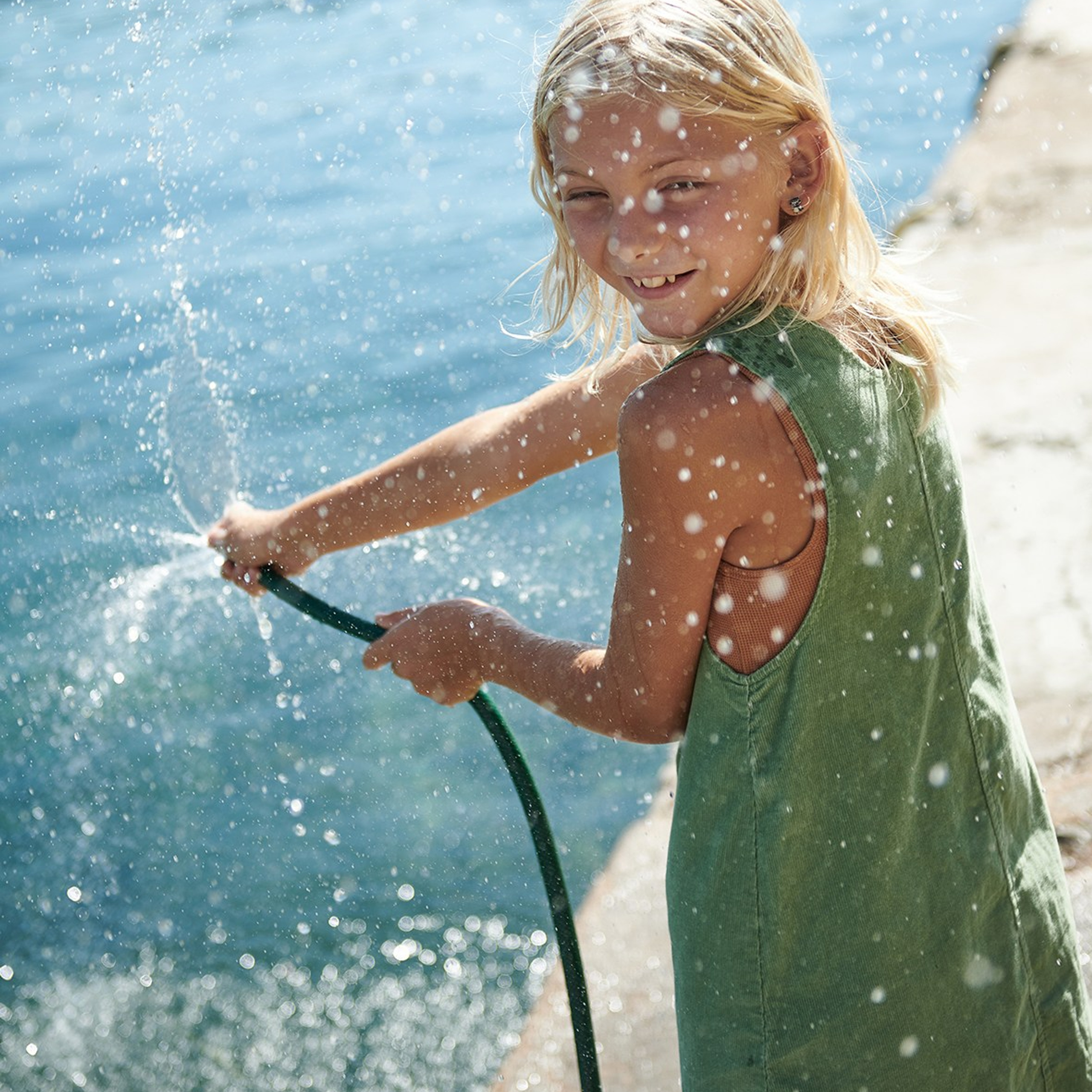 Playful portrait of a blonde girl in a green dress splashing water during a children’s photoshoot