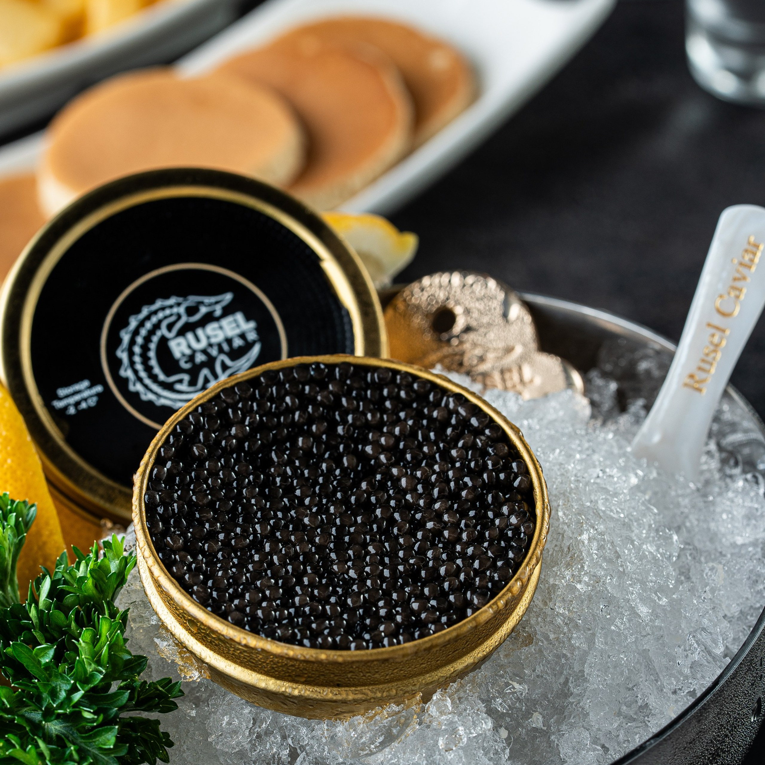 Buz üzərində açılmış Rusel Caviar qara kürüsü, ağ qaşıqla birlikdə, fon planda kərə yağı dilimləri və mini blinçiklər.
