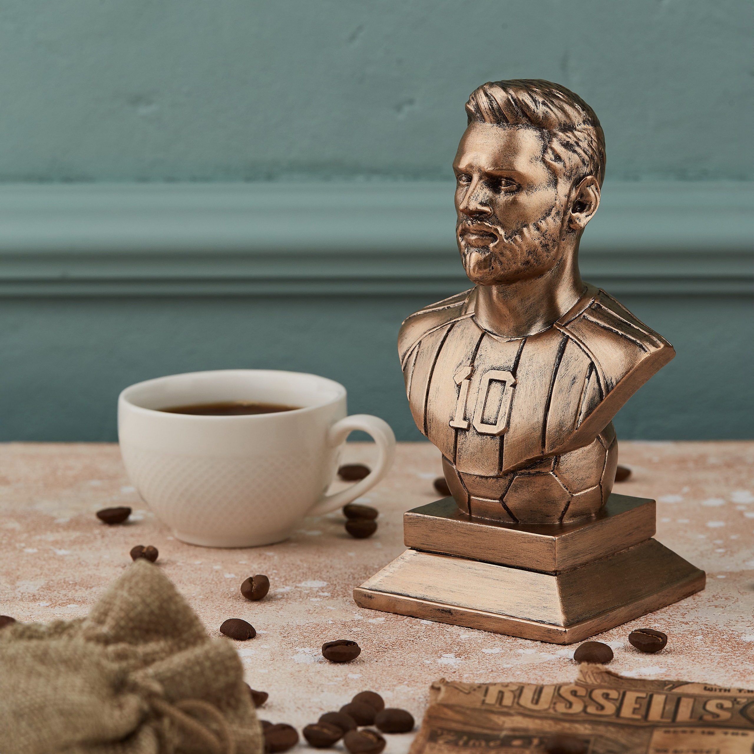 Lionel Messinin bürünc büst heykəli, səhər dekorasiyası: fincan qəhvə, qəhvə dənələri və qəzet yanında.