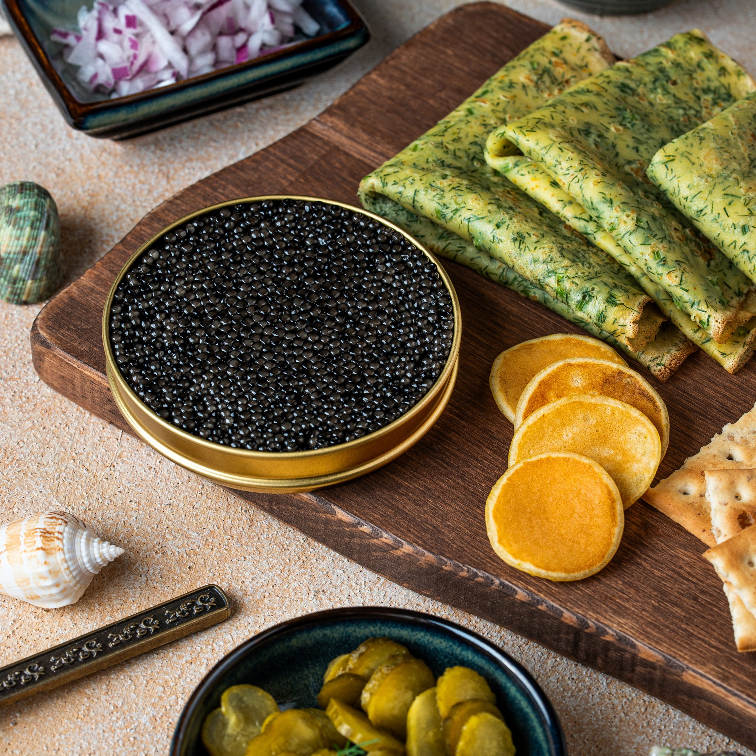 Qara kürülü metal banka, yanında şüyüdlü blinçiklər, mini blinlər, turşu xiyar və doğranmış qırmızı soğan ilə süfrə kompozisiyası.