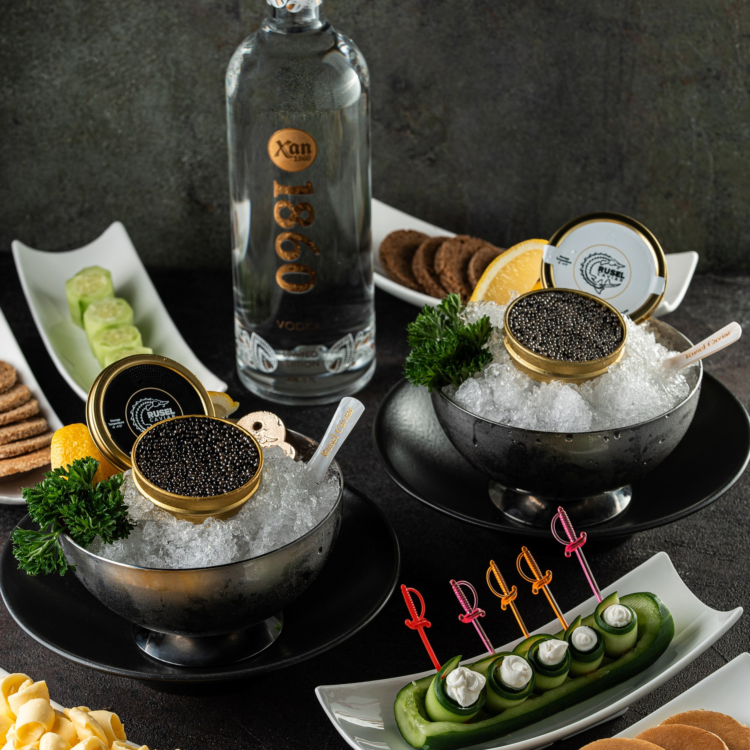 Zəngin süfrə düzümü: Rusel Caviar qara kürüsü iki qabda buz üzərində, mərkəzdə Xan 1860 votkası və ətrafda müxtəlif qəlyanaltılar və blinçiklər.