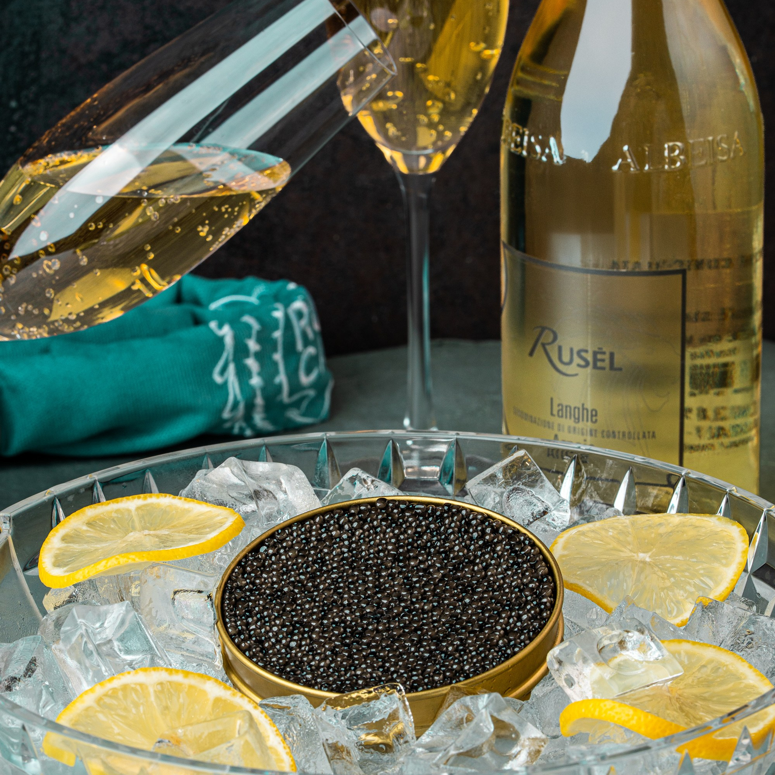 Şüşə qabda buzların üzərində limon dilimləri ilə təqdim olunmuş qara kürü və arxa planda Rusel şərabı və şampan qədəhləri.