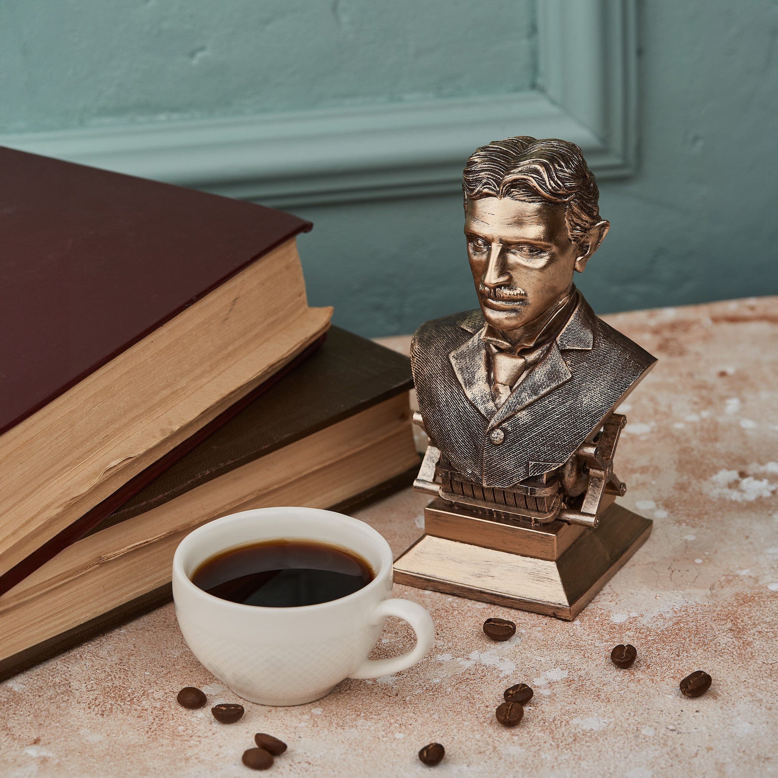 Nikola Teslanın bürünc büst heykəli, iki köhnə kitabın və fincan qəhvənin yanında, elmi bəzək kimi.