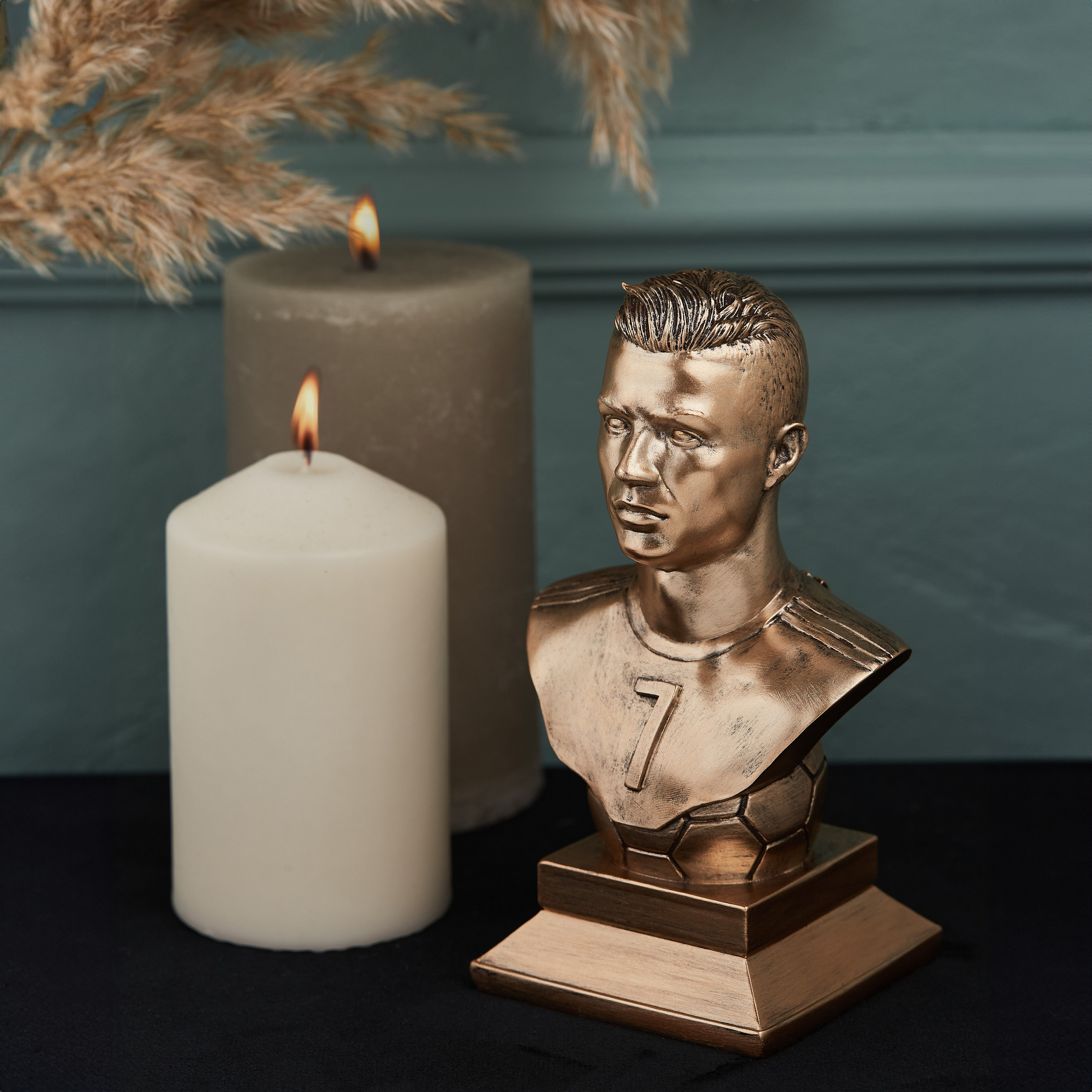 Kriştiano Ronaldonun bürünc büst heykəli, yandırılmış şamlar və quru ot (pampas otu) fonunda masa dekorasiyası.