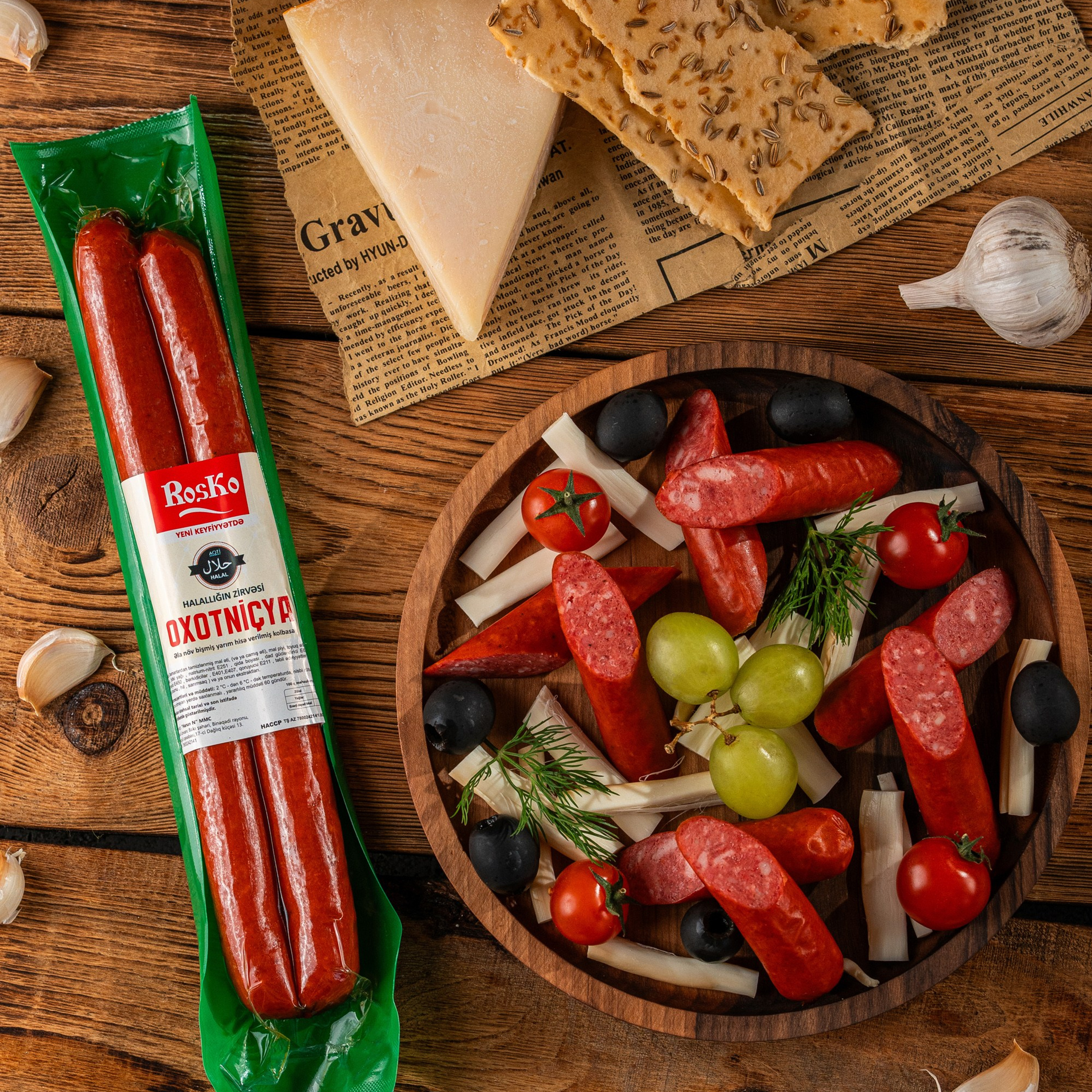 Roska “Oxotniçya” kolbasası paketli və dilimlənmiş formada, pendir, kraker, zeytun və üzüm ilə rustik taxta fon üzərində təqdim olunub.