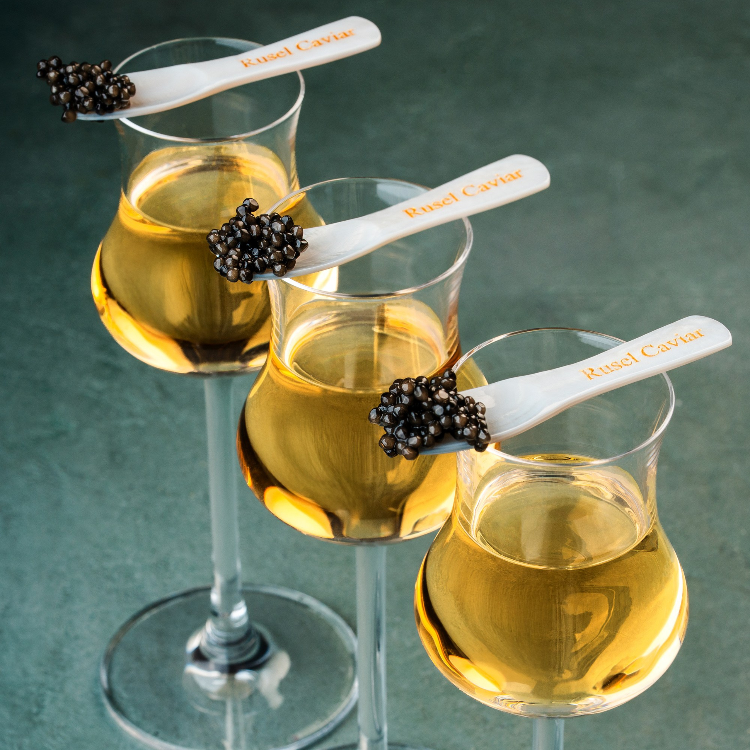 Zərif dizaynda şərab qədəhləri üzərində incə qaşıqlarda təqdim olunan qara kürü — "Rusel Caviar" brendi ilə dekor edilmiş vizual.