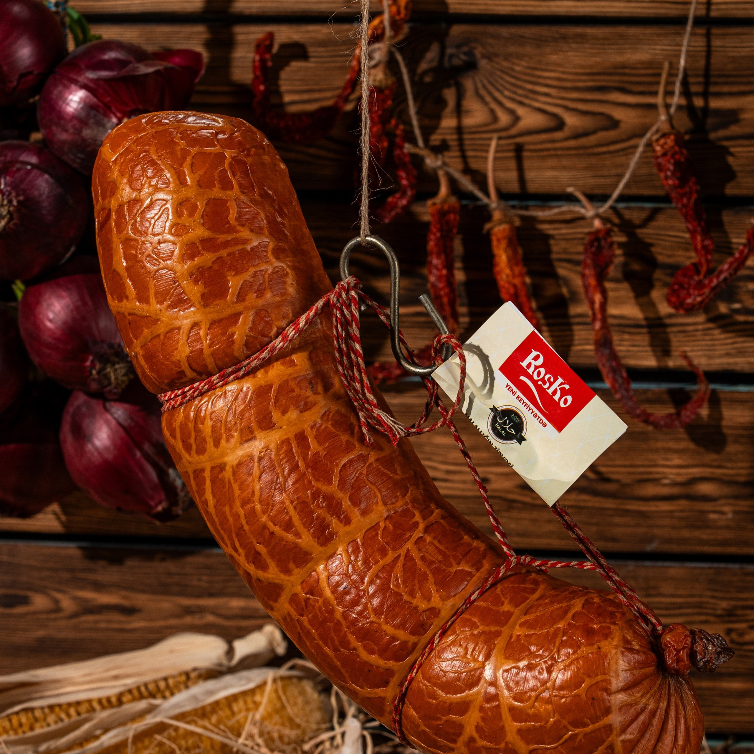 Roska kolbasası kənd üslubunda asılmış formada, taxta fon və qurudulmuş ədviyyatlarla birlikdə rustik atmosferdə təqdim olunub.