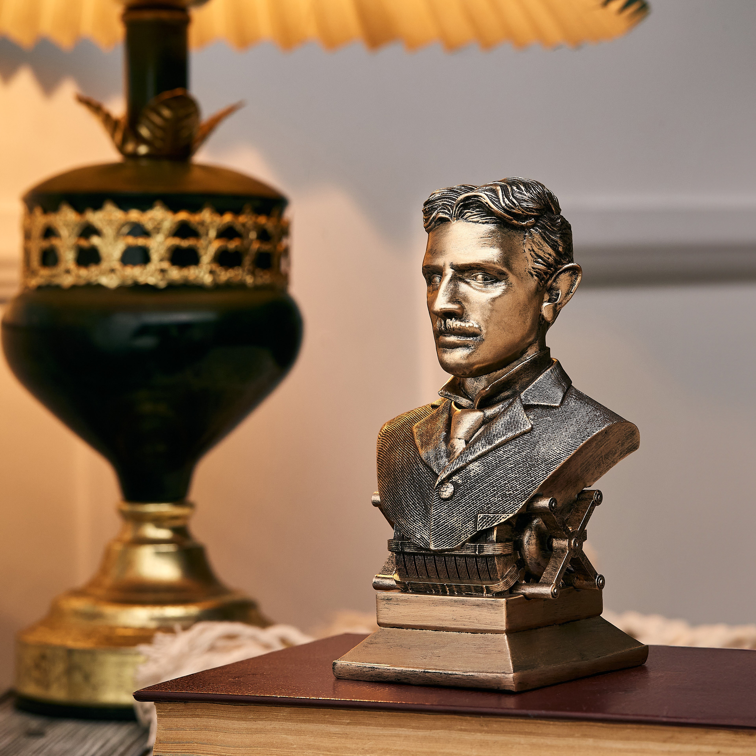 Nikola Teslanın bürünc büstü, köhnə kitabın üzərində, lampa və yumşaq işıqlandırma ilə bəzədilmiş masa dekorasiyası.