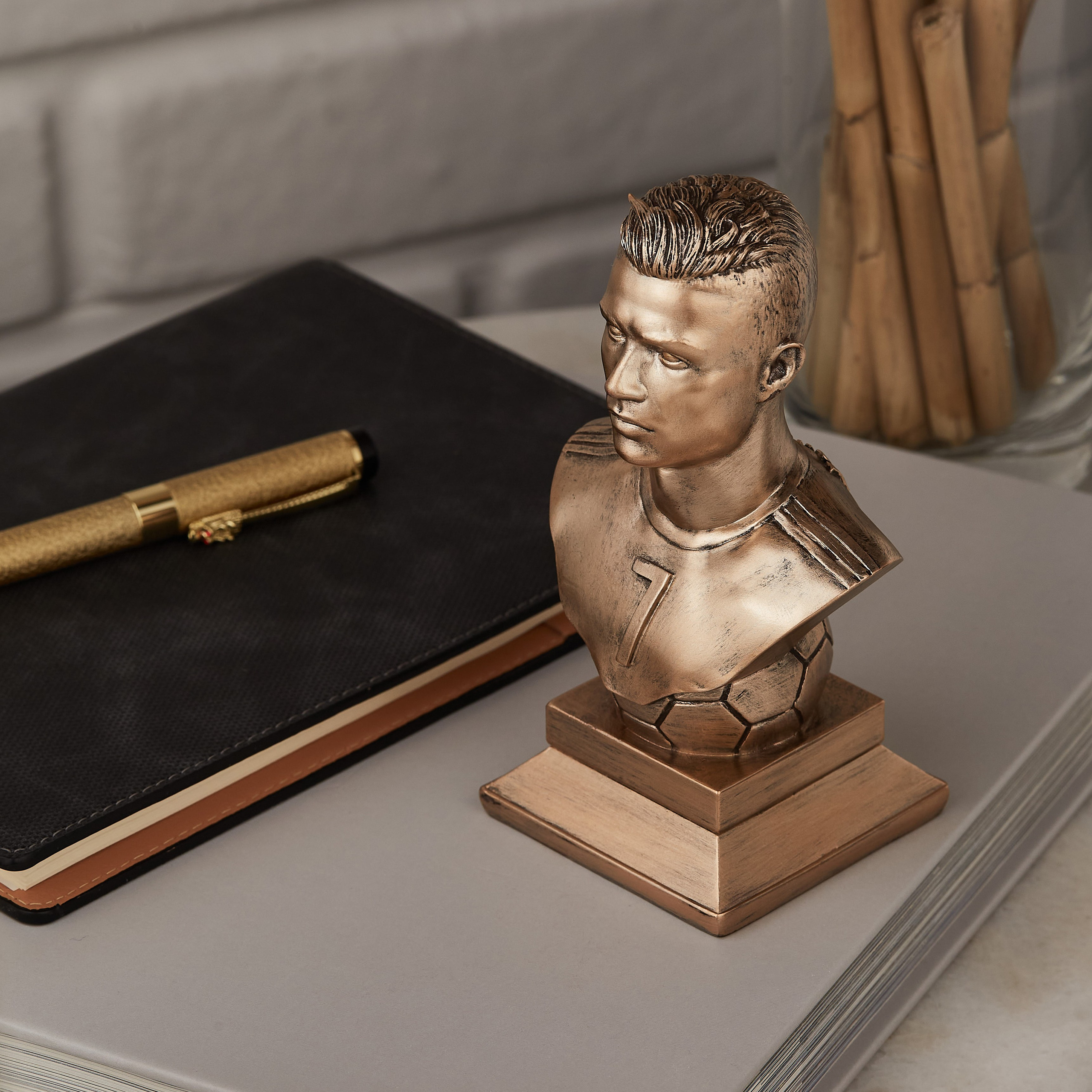 Kriştiano Ronaldonun bürünc rəngli büst heykəli, 7 nömrəli forması ilə, dəftər və qızılı qələm yanında masa dekorasiyası.