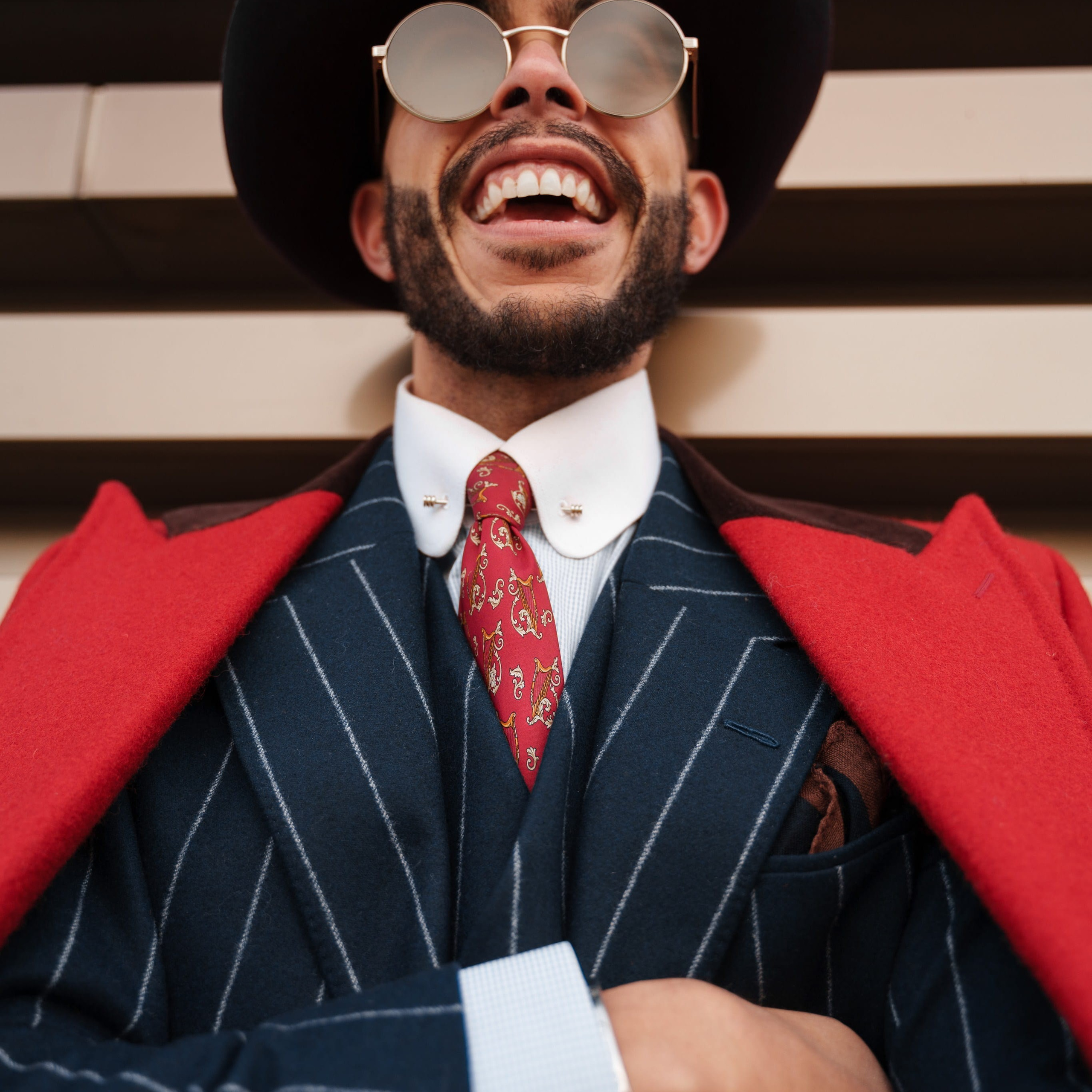 A man in a dapper pinstriped suit and bold red coat laughs heartily, his joy amplified by round sunglasses and a stylish fedora, creating a lively and charismatic portrait.