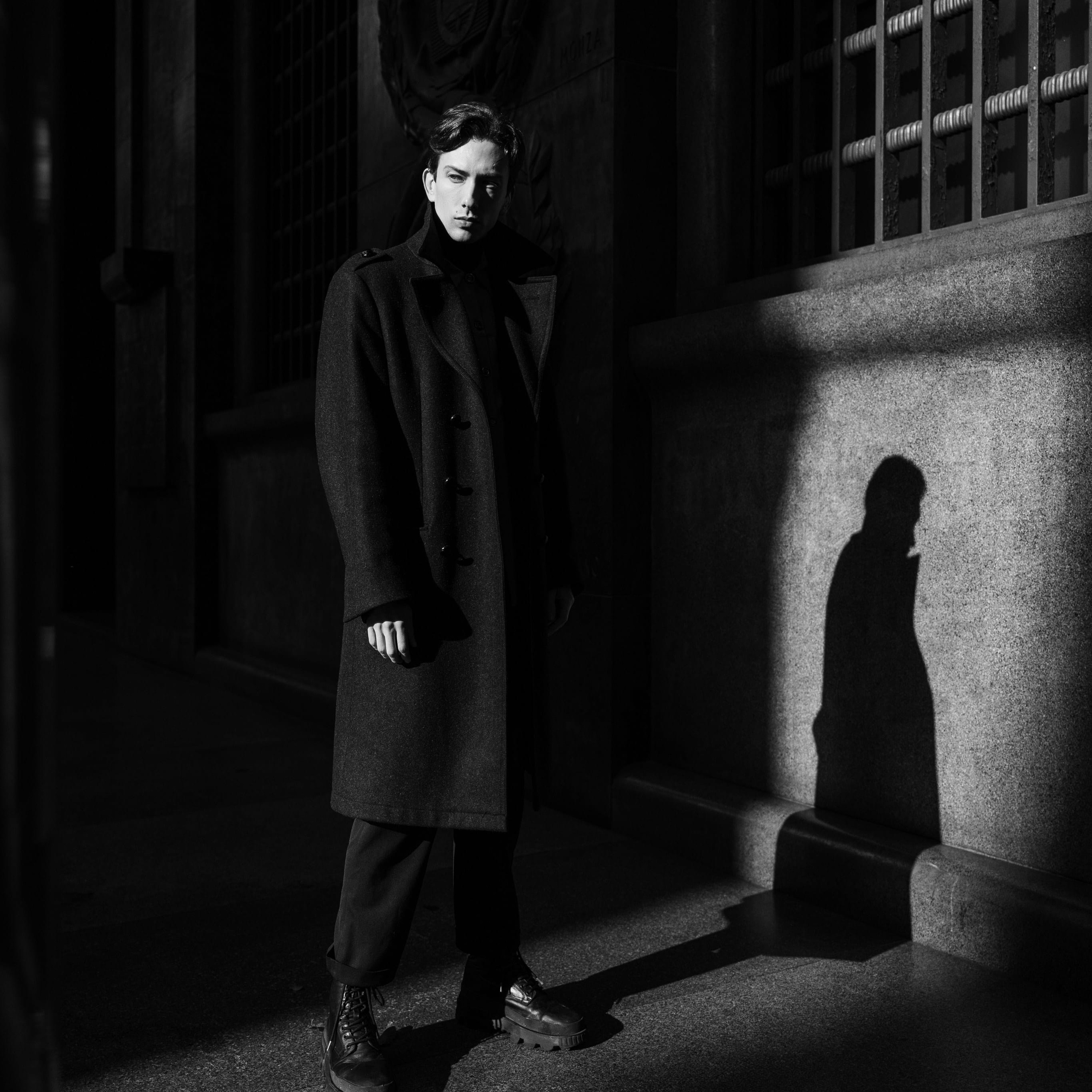 Dramatic black and white portrait of a man in a long coat casting a shadow on a stone wall in Milan