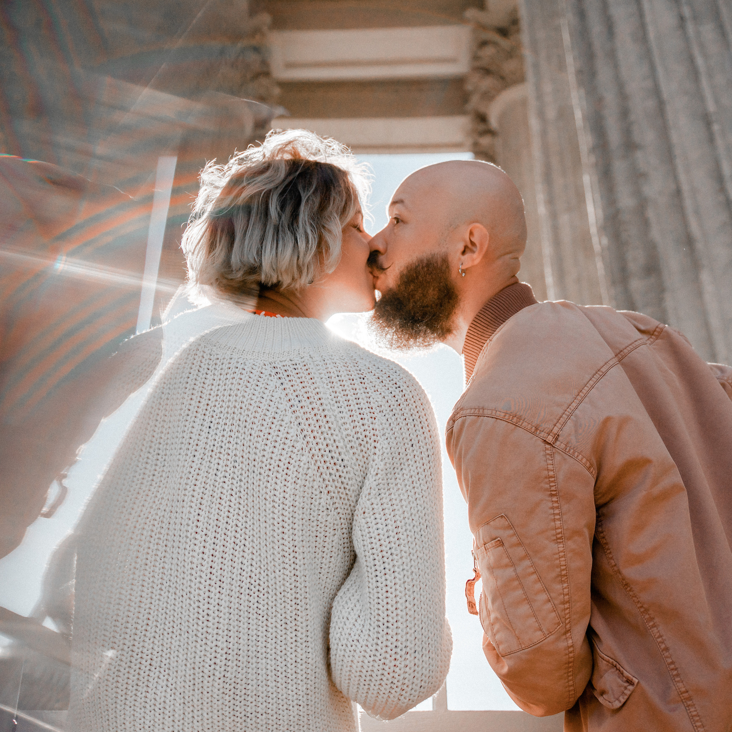 Romantic backlit couple kissing under historic architecture, with lens flare and chromatic aberration adding dreamy atmosphere, shot with a prime lens, possibly 50mm, for crisp subject detail against a soft background
