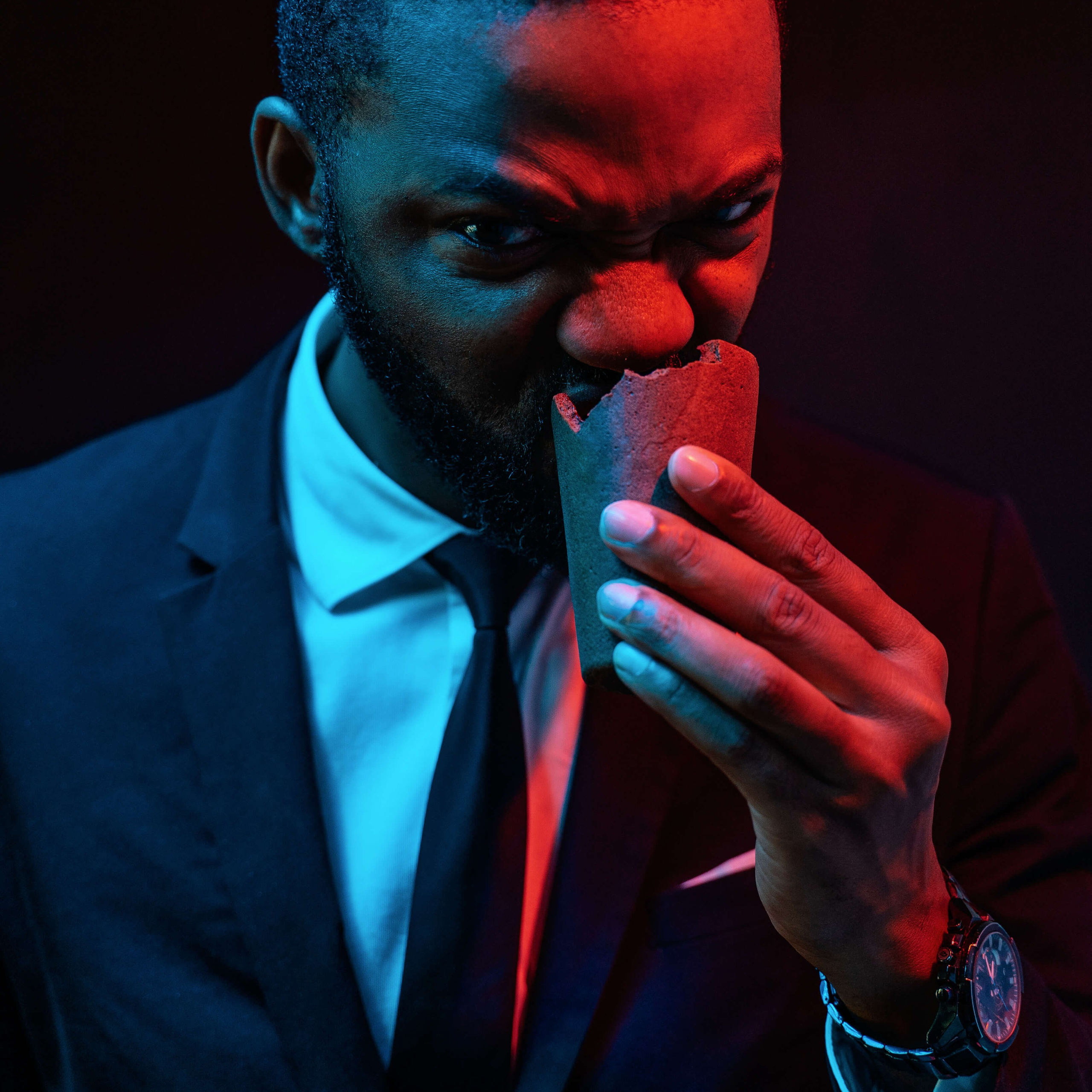 A man in a sharp suit exudes mystery and intensity as he bites into an ice cream sandwich, illuminated by dramatic red and blue lighting that casts a cinematic glow on the scene.