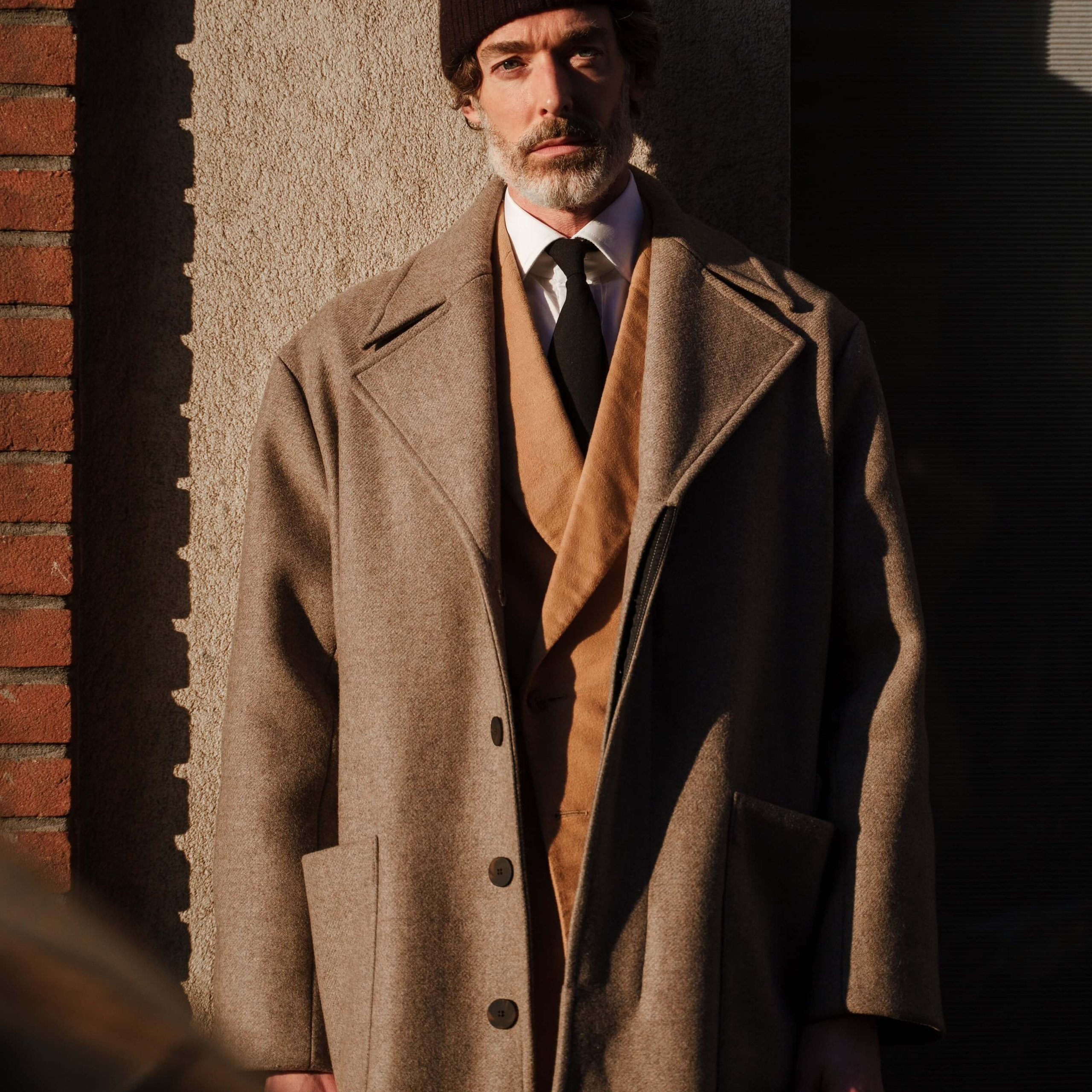 Shot of a thoughtful man dressed in timeless attire, with sunlight casting sharp shadows, highlighting a blend of urban fashion and natural light.