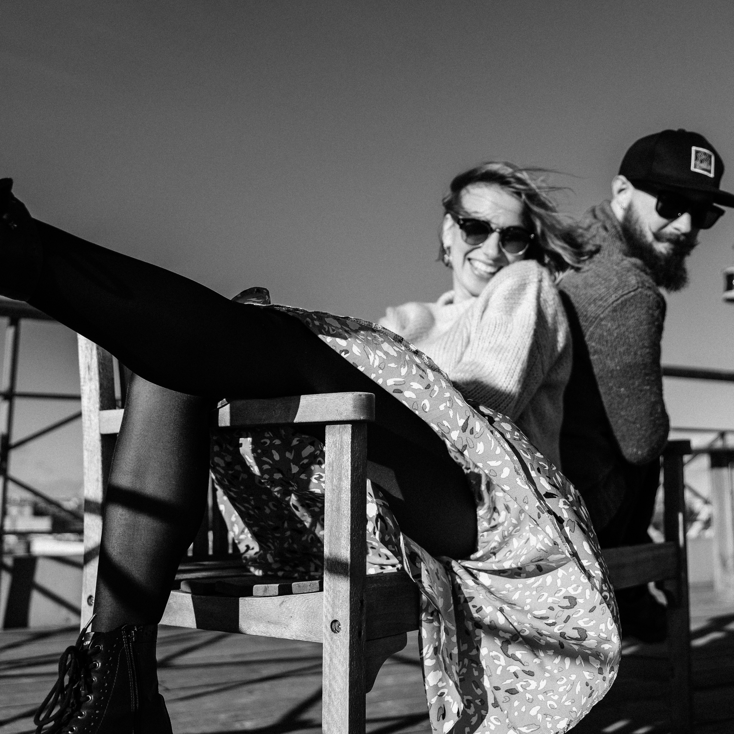 Dynamic black and white candid shot of couple laughing on rooftop, with focus on woman's playfully kicked-up foot; possibly captured with a 35mm prime lens for its storytelling field of view