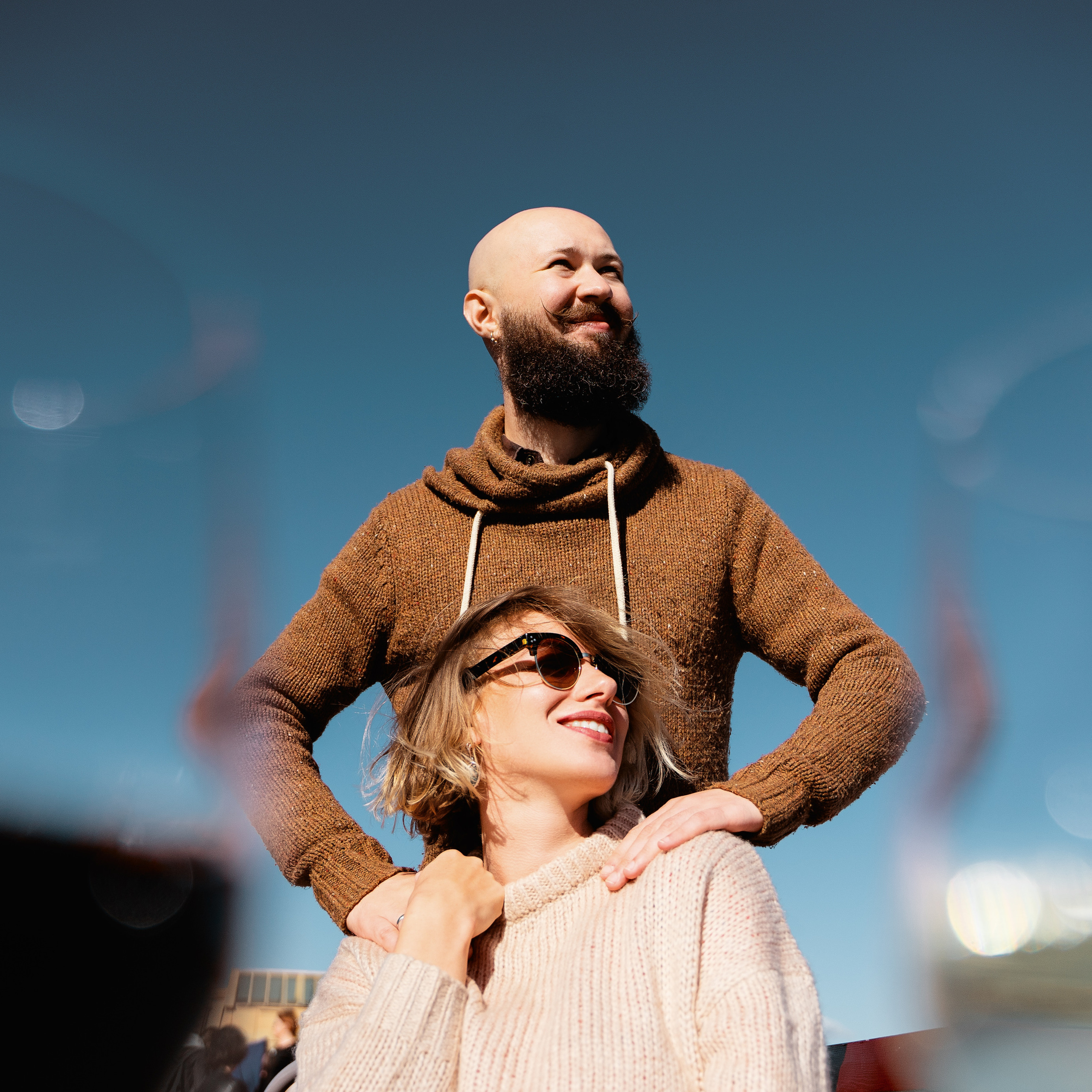 Low angle portrait of couple with clear blue sky, utilizing a bright lens flare for artistic effect, suggesting use of a large aperture lens, such as a 50mm f/1.4, for depth and bokeh.