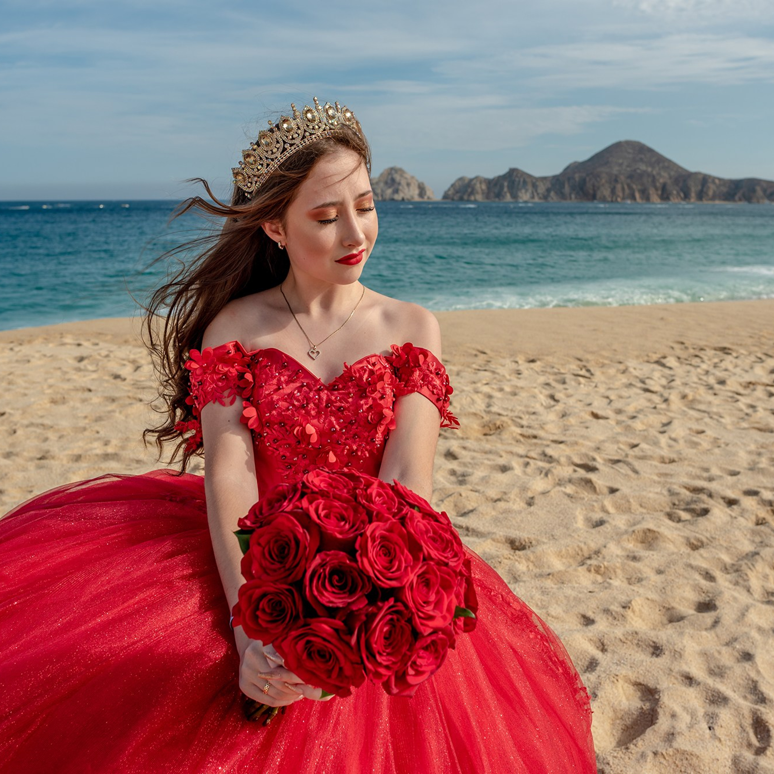 XV años photoshoot in Cabo San Lucas – Sofie wearing a stunning red quinceañera dress with a bouquet of red roses, photographed at El Médano beach with the Finisterra rock formations in the background. Destination XV photographer in Los Cabos