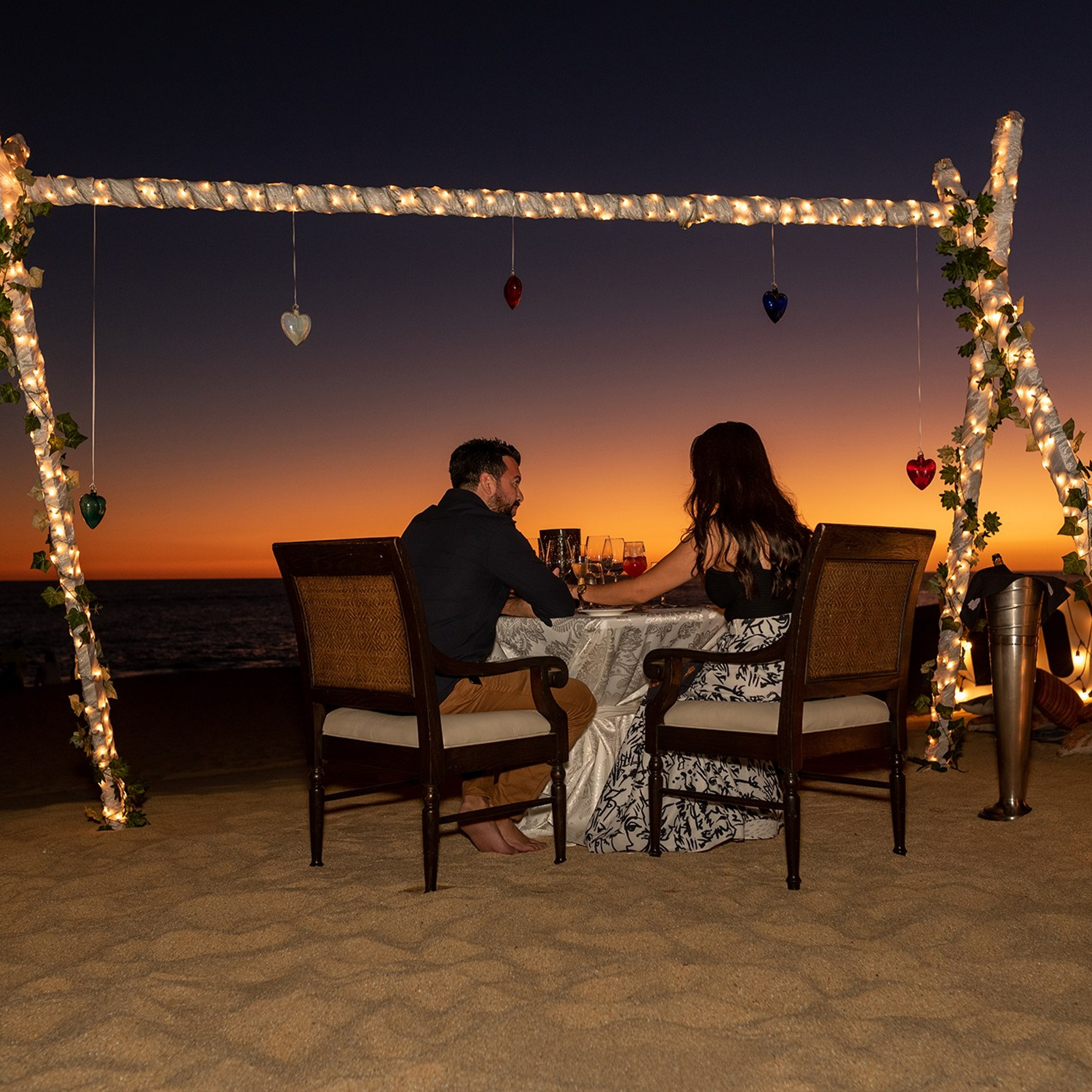 Cabo San Lucas engagement photography – candid moment of newly engaged couple sitting in a romantic setup with fairy lights, back to the camera, enjoying the orange sunset after their proposal