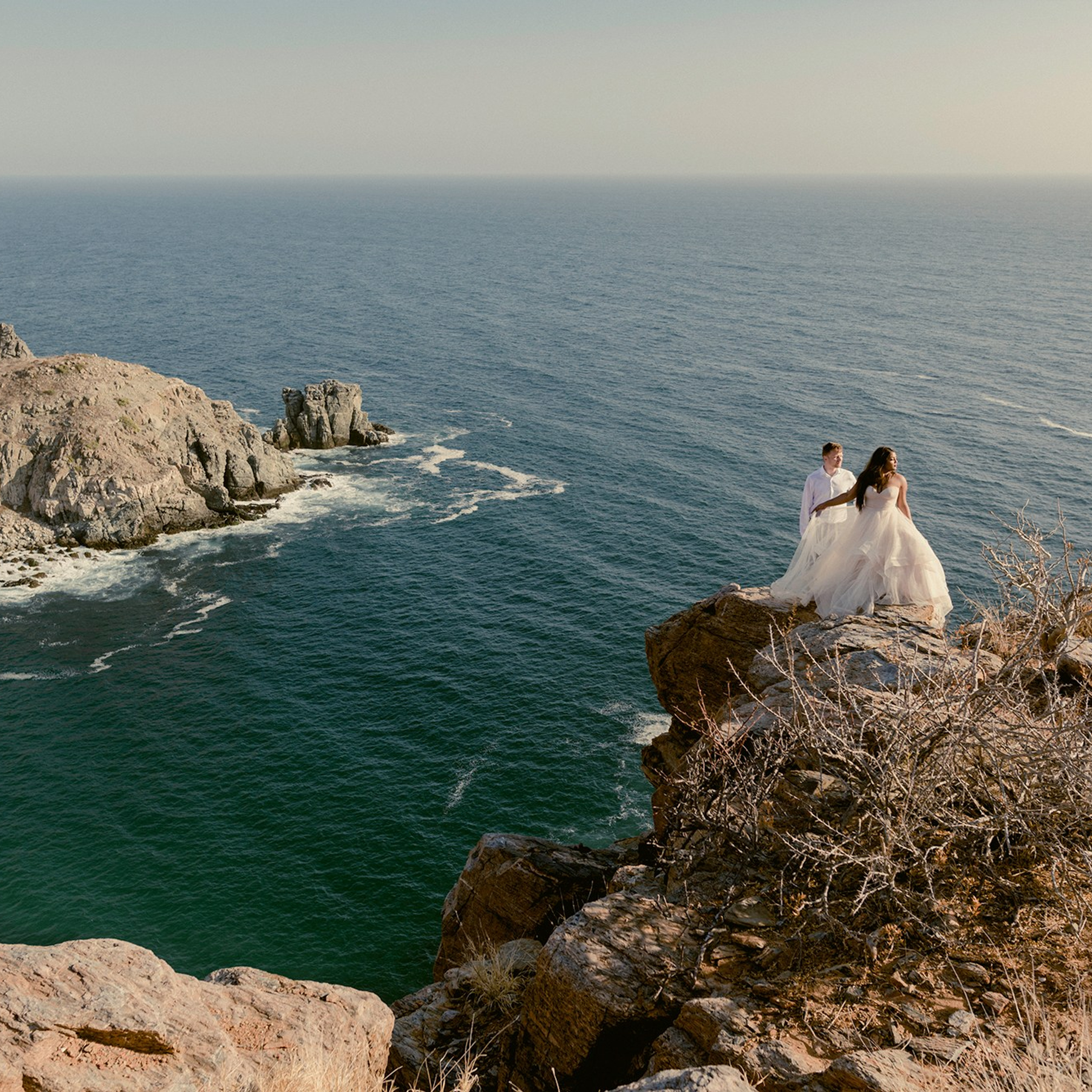 Trash the dress photoshoot in Los Cabos; bride and groom embracing on coastal rocks with ocean waves and desert scenery, captured by a destination wedding photographer in Baja California Sur