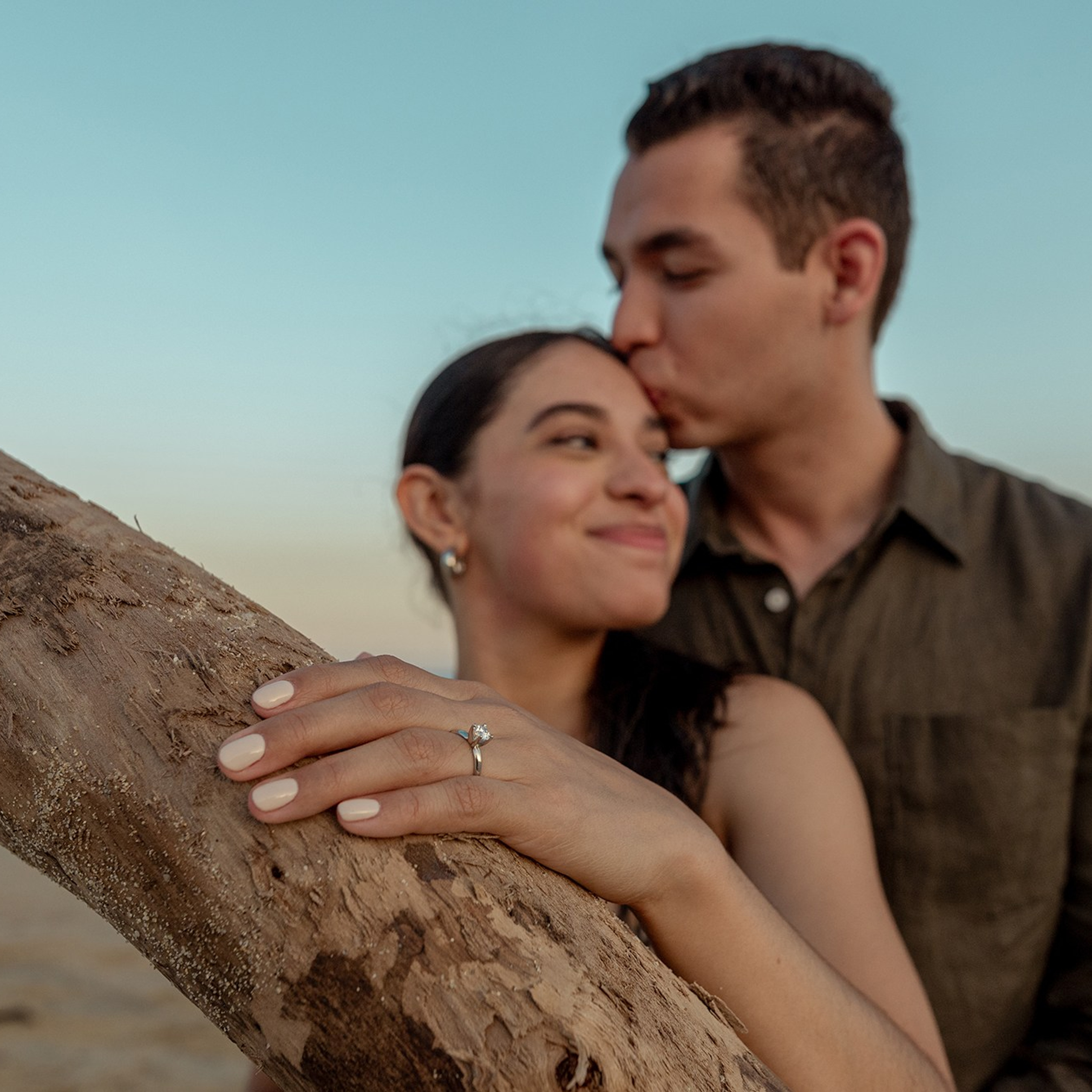 Engagement photoshoot in Los Cabos – close-up of bride-to-be’s hand showing the engagement ring, while fiancé embraces her from behind and kisses her forehead at sunset by the ocean