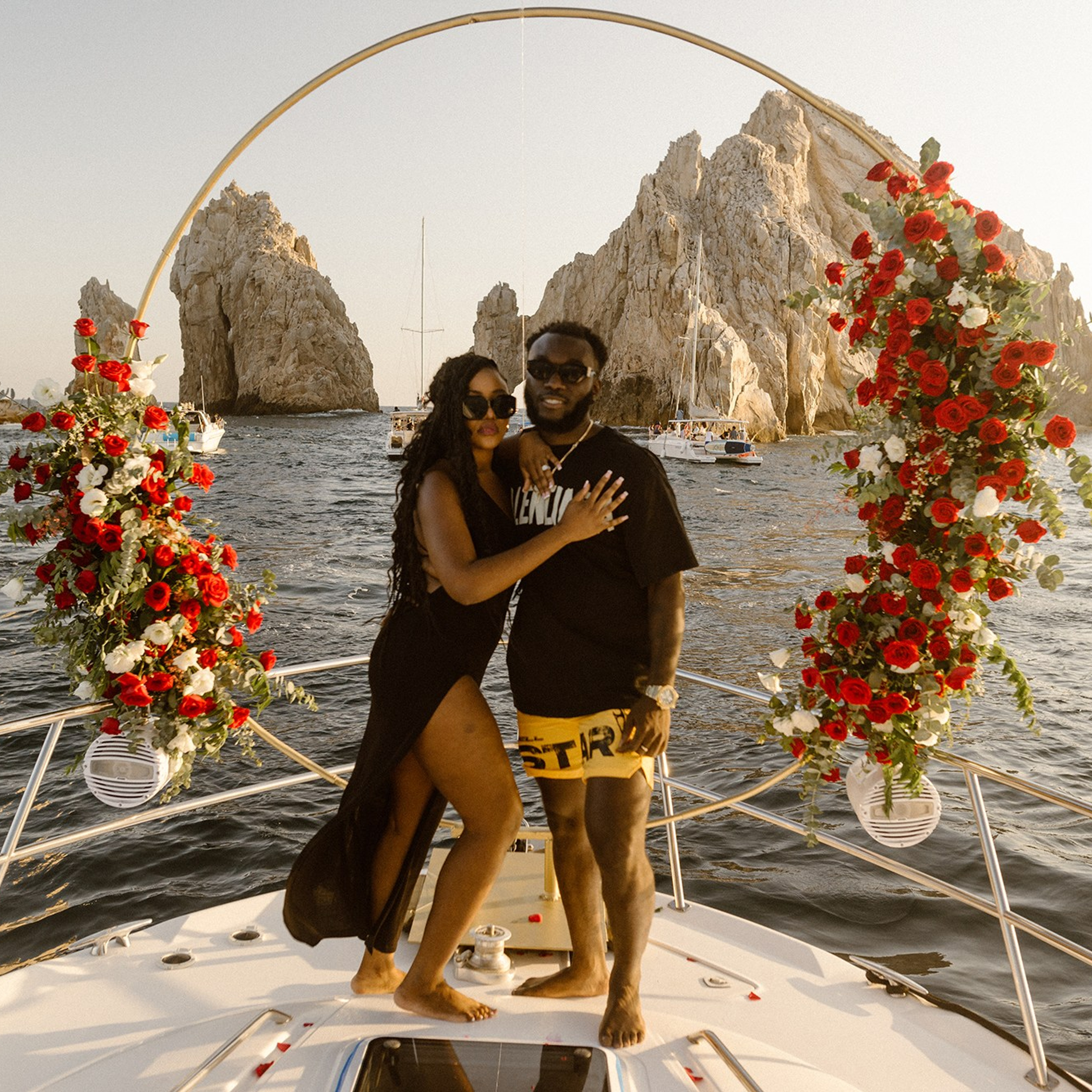 Romantic engagement photography in Cabo San Lucas – Afro couple posing on a yacht with luxury red floral arrangement, El Arco de Los Cabos in the background, destination photographer for proposals, engagements and couples in Los Cabos.