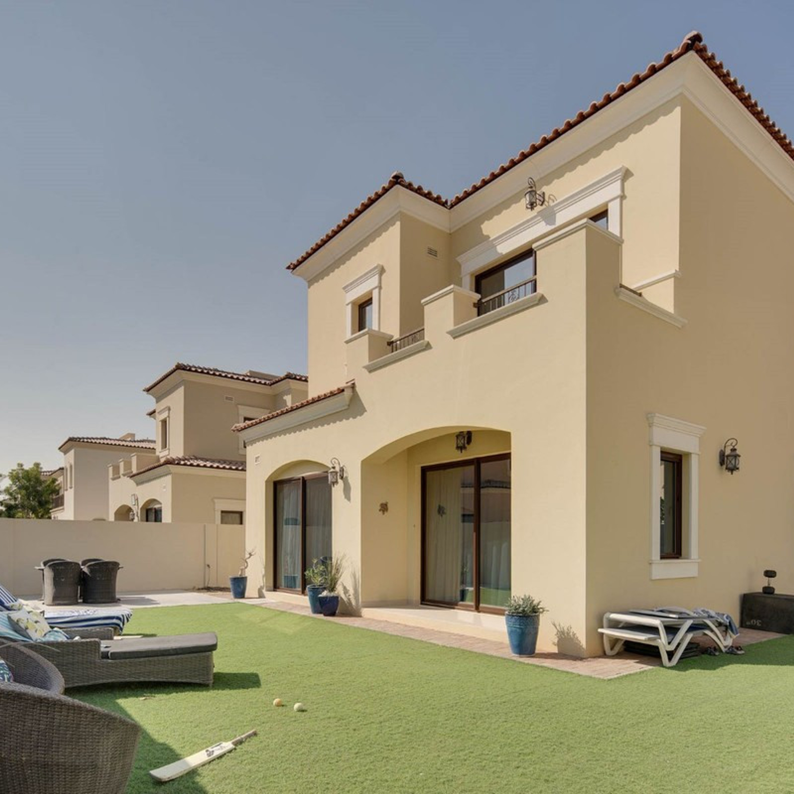 Two-story beige villa with a red tile roof, featuring an artificial grass yard and outdoor lounge chairs