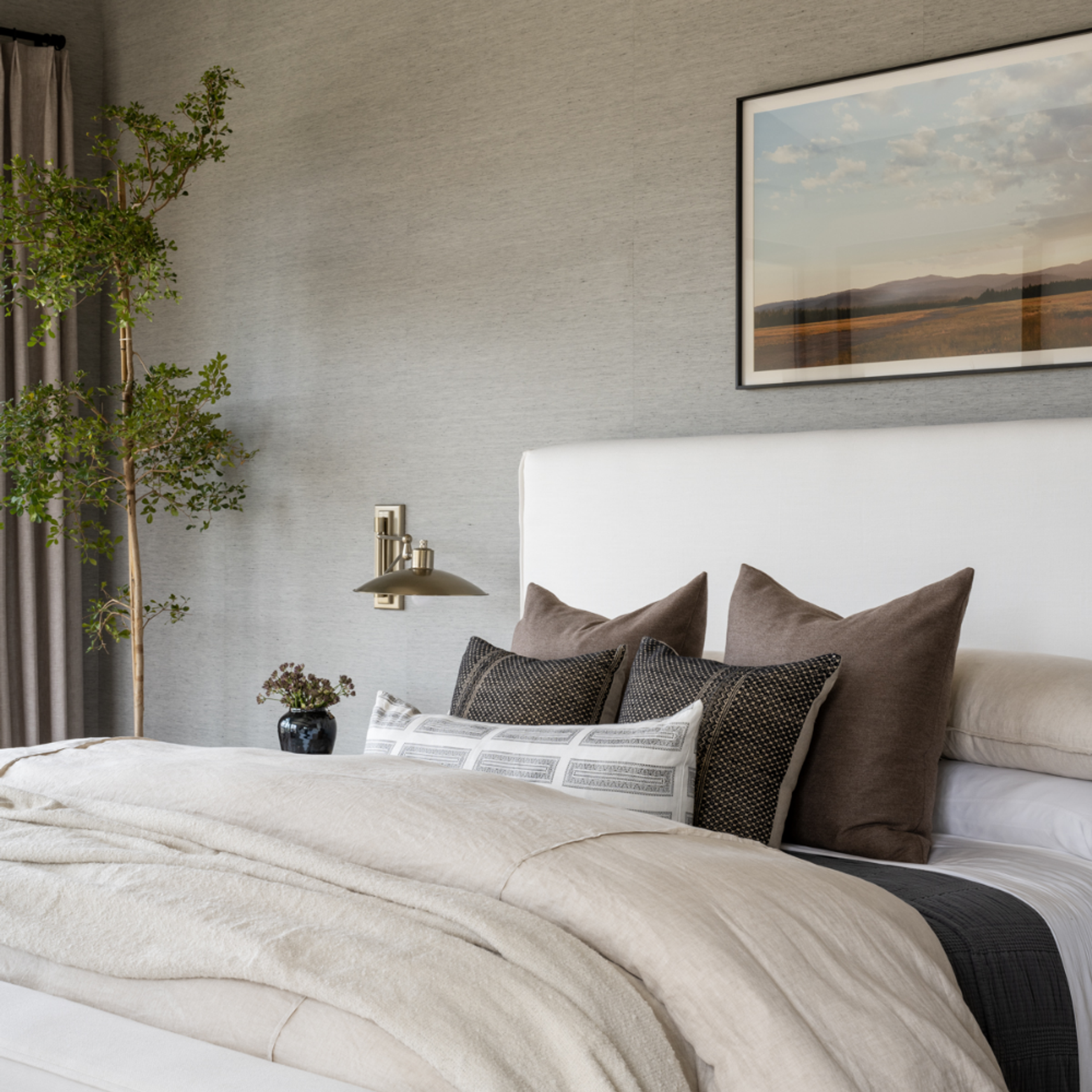 Stylish bedroom interior with neutral-toned bedding, a wall-mounted light fixture, and a framed landscape photograph above the bed