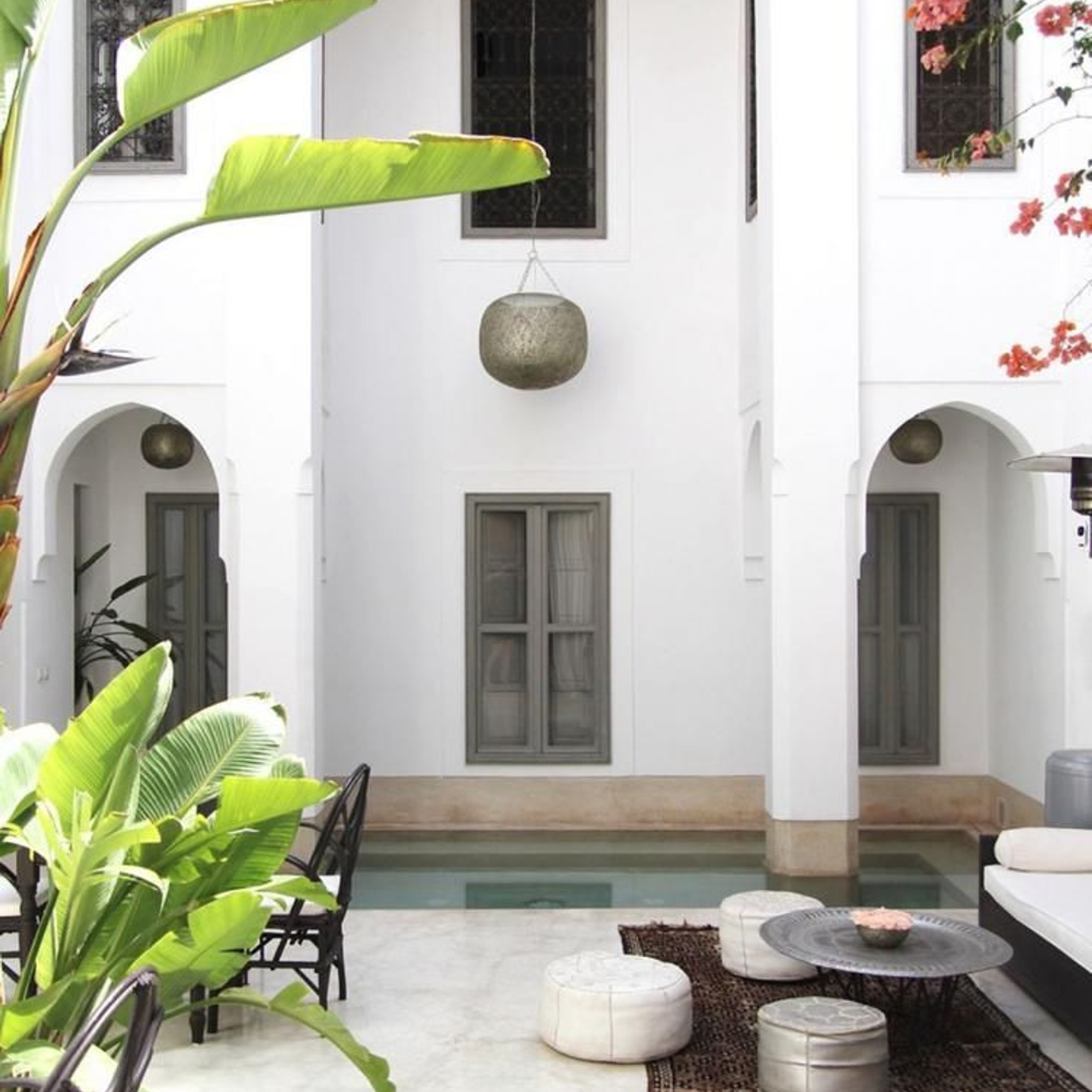 Bright, open courtyard with white walls, tropical plants, and a small pool, featuring minimal seating on a rug, poufs, and an elegant hanging light