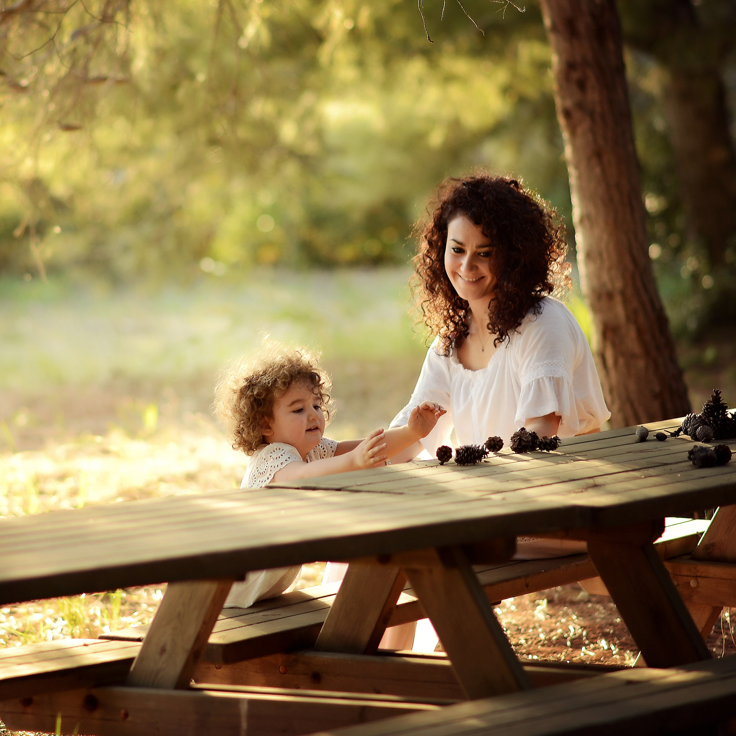 A mother and daughter playing with pinecones on a wooden table in the forest, bathed in soft, dreamy light.