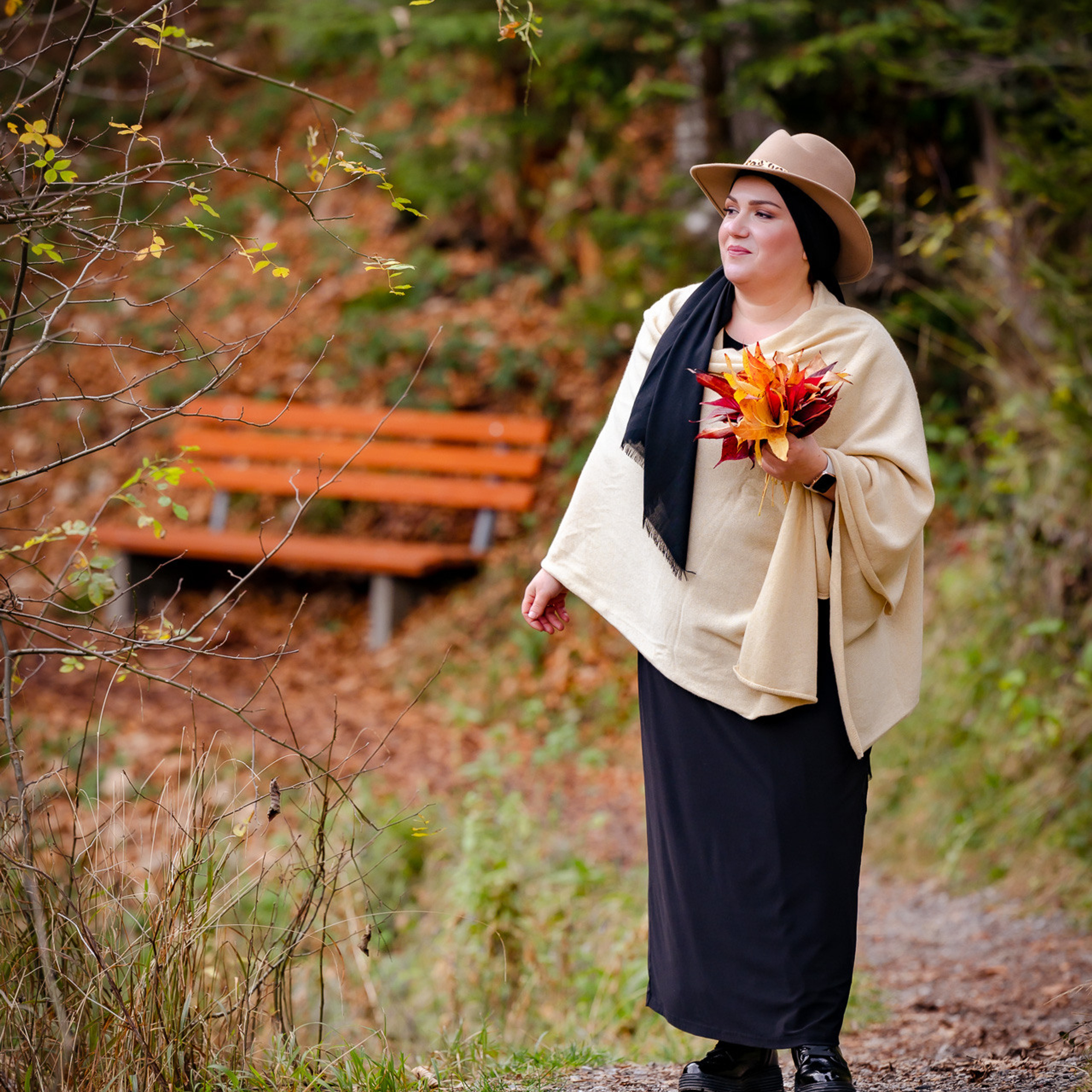 Eine Frau in einem beigen Umhang und Hut mit einem Strauß Herbstblättern in der Hand spaziert entlang einer orangefarbenen Bank.