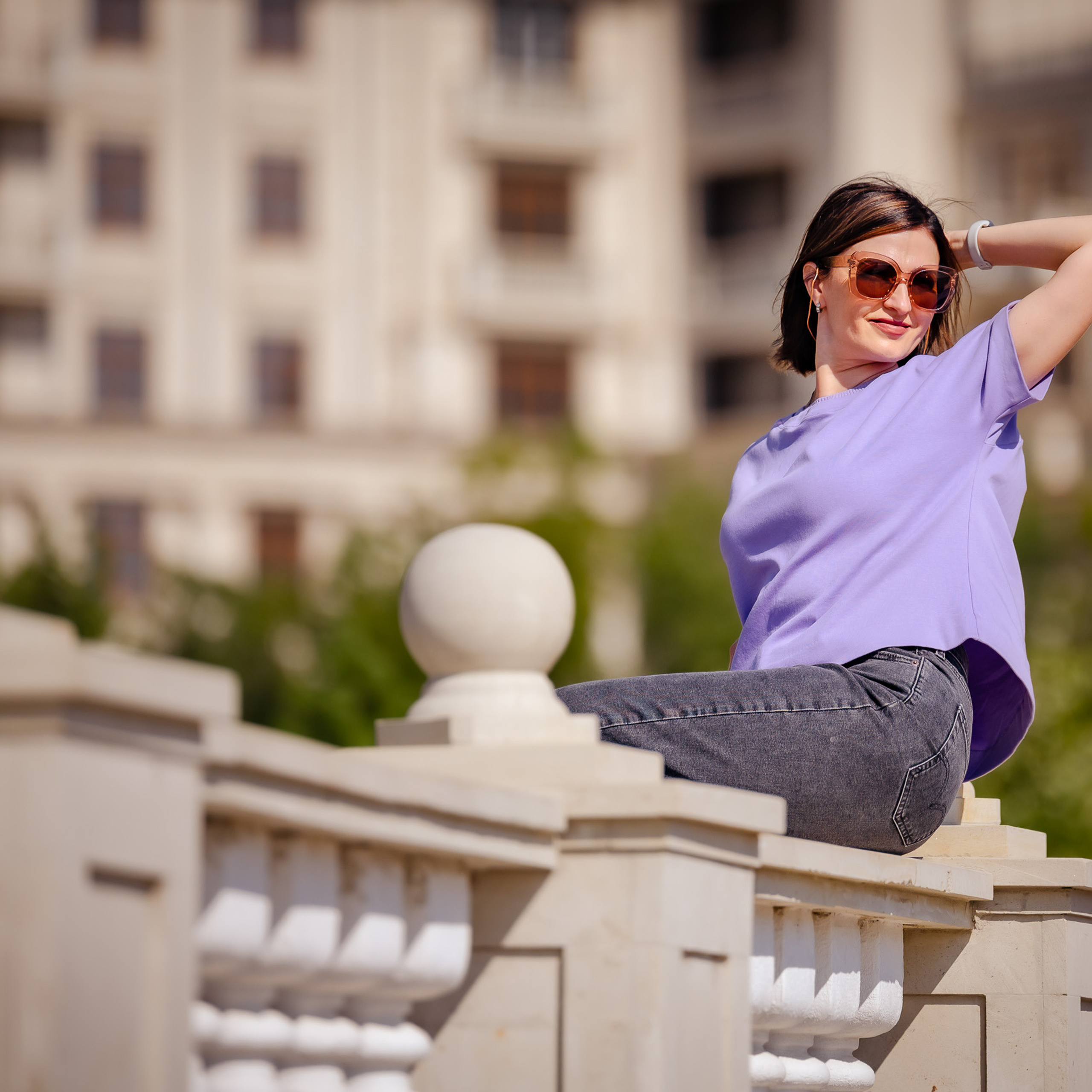 Ein Mädchen in einem T-Shirt und Jeans sitzt auf einer steinernen Balustrade entlang einer städtischen Allee.